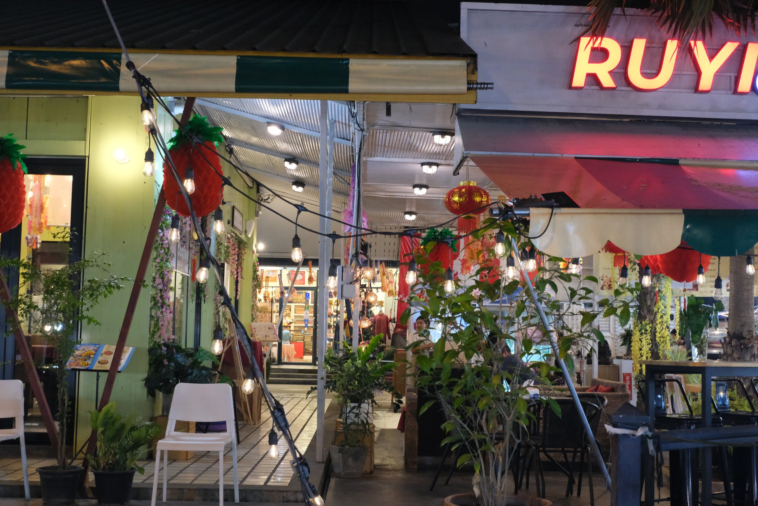 Nighttime view of a restaurant or cafe with outdoor seating, hanging string lights, potted plants, decorative red lanterns, and a bright red sign with partially visible letters.