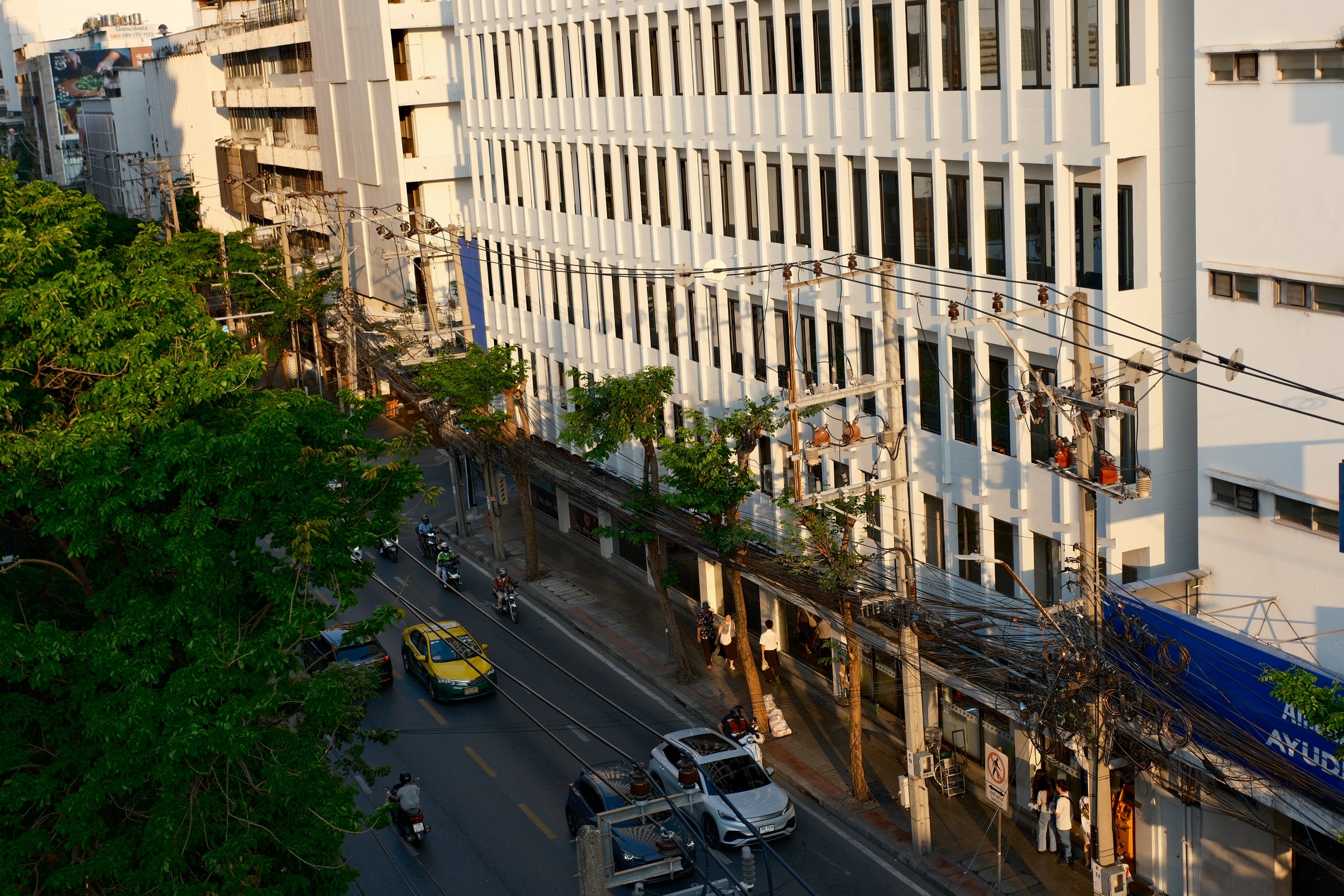 City street scene with cars and motorcycles, pedestrians walking on the sidewalk, a large white modern building with vertical window panels, and multiple power lines crossing the street.