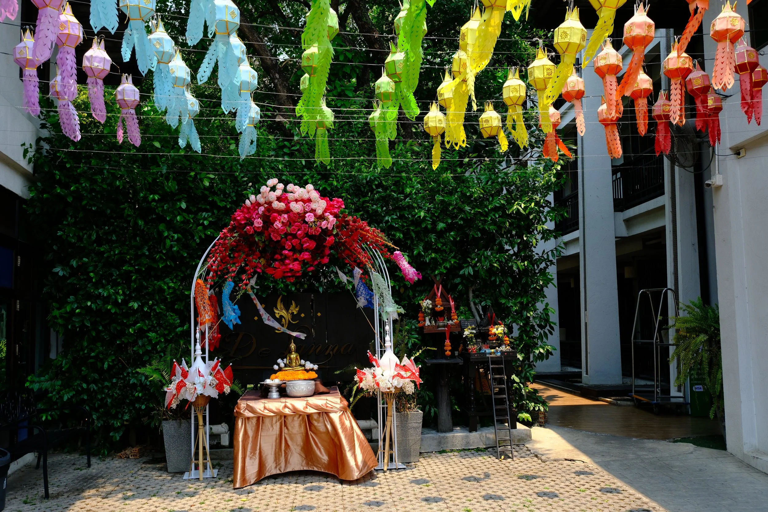 Outdoor setup with a floral arrangement and an altar with Buddhist statues, surrounded by colorful paper lanterns hanging overhead in a garden area.