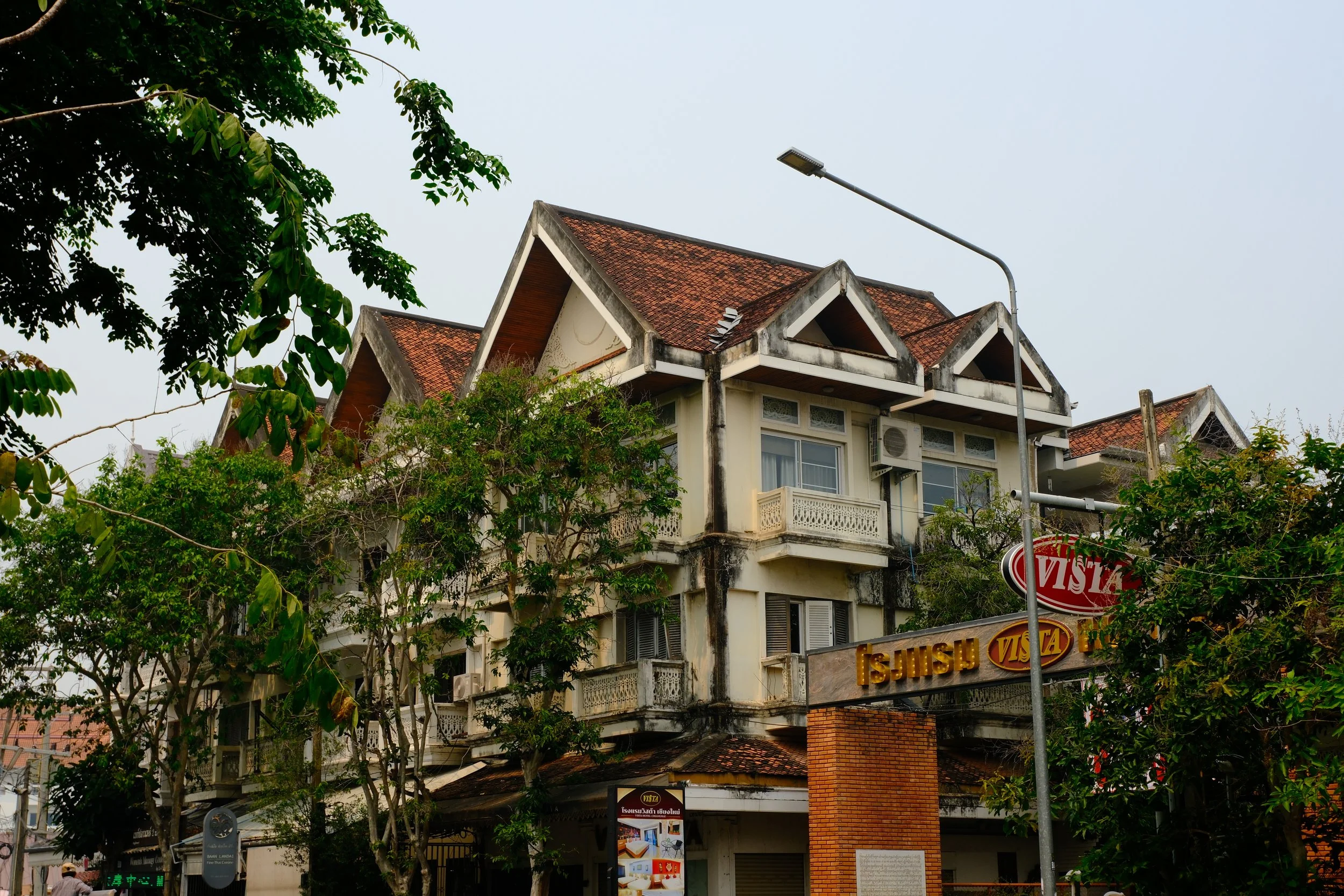 An old multi-story building with a sloped red-tile roof, blackened and weathered walls, surrounded by trees and street signs, with a cloudy sky in the background.