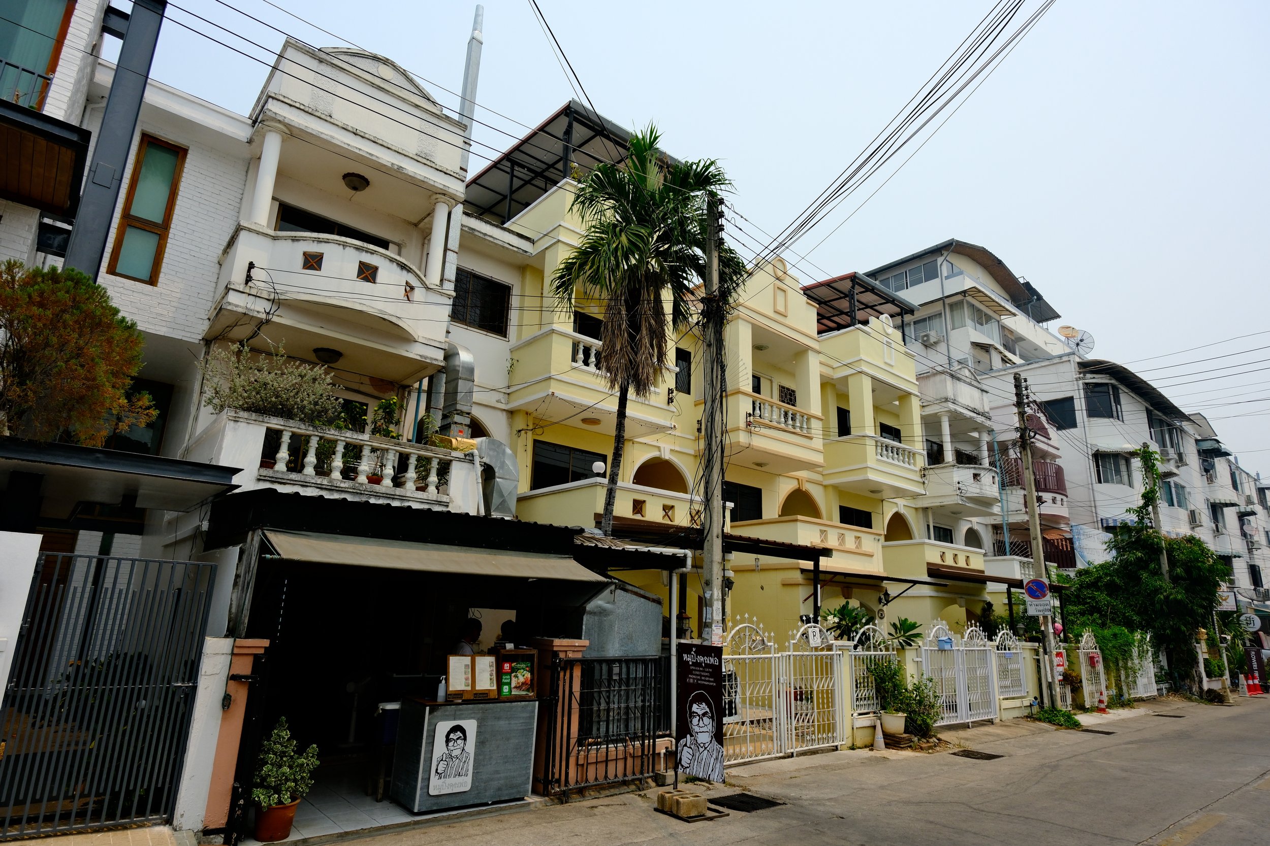 Multiple multi-story residential buildings with balconies, surrounded by fences and gate, along a street with power lines and trees.