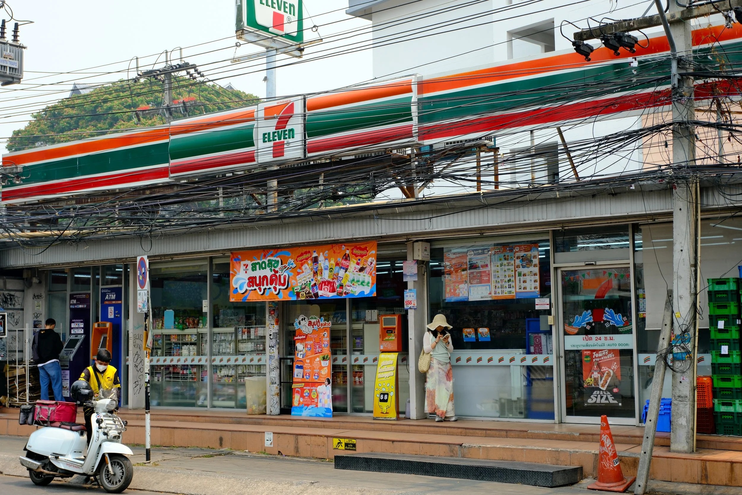 A 7-Eleven convenience store on a street with people and a scooter in front, a woman wearing a hat and mask standing outside, electrical wires above, and signage in a foreign language.