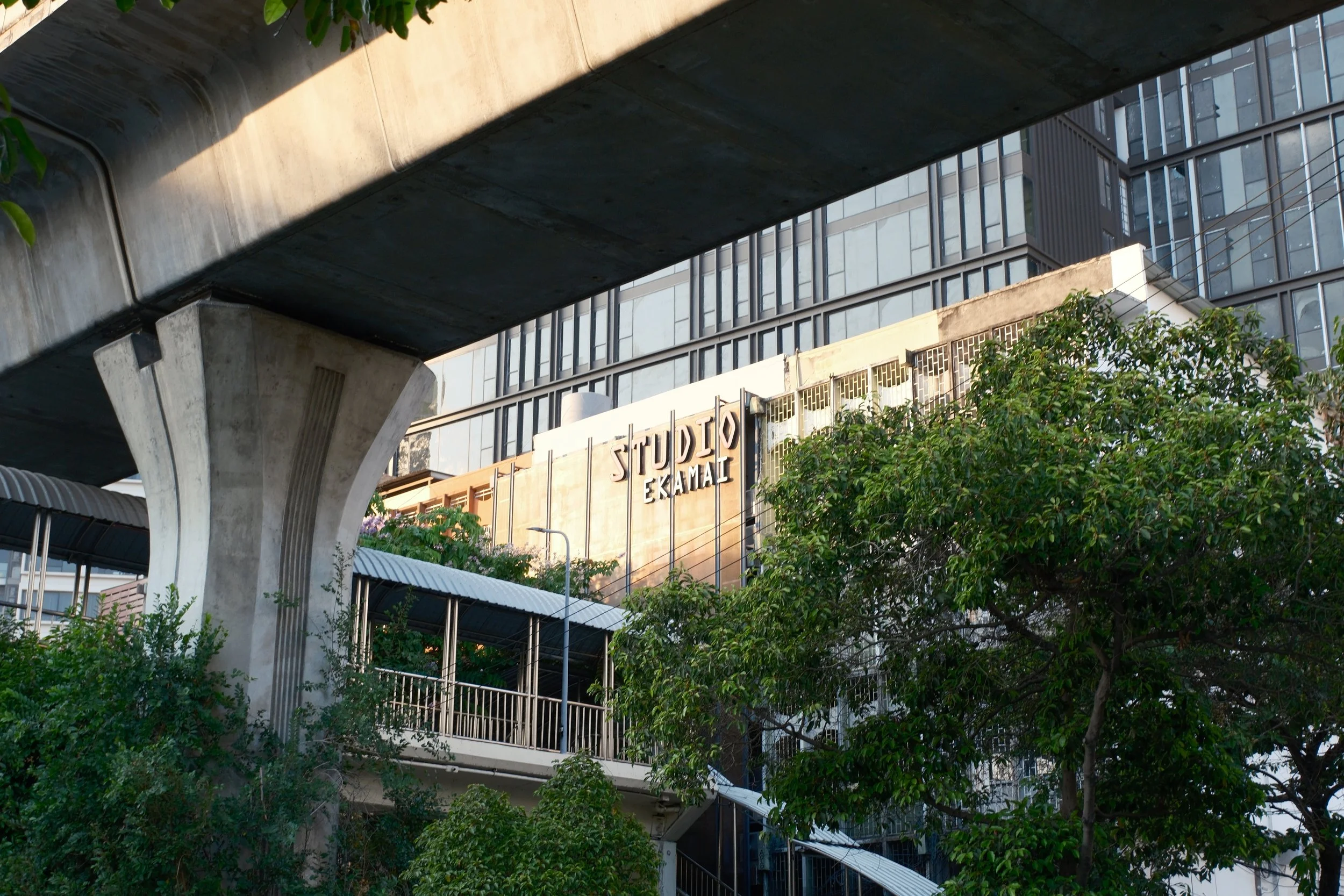 View of a cityscape with trees, a pedestrian bridge, and a building with a sign that reads 'Studio Ekkamai.'