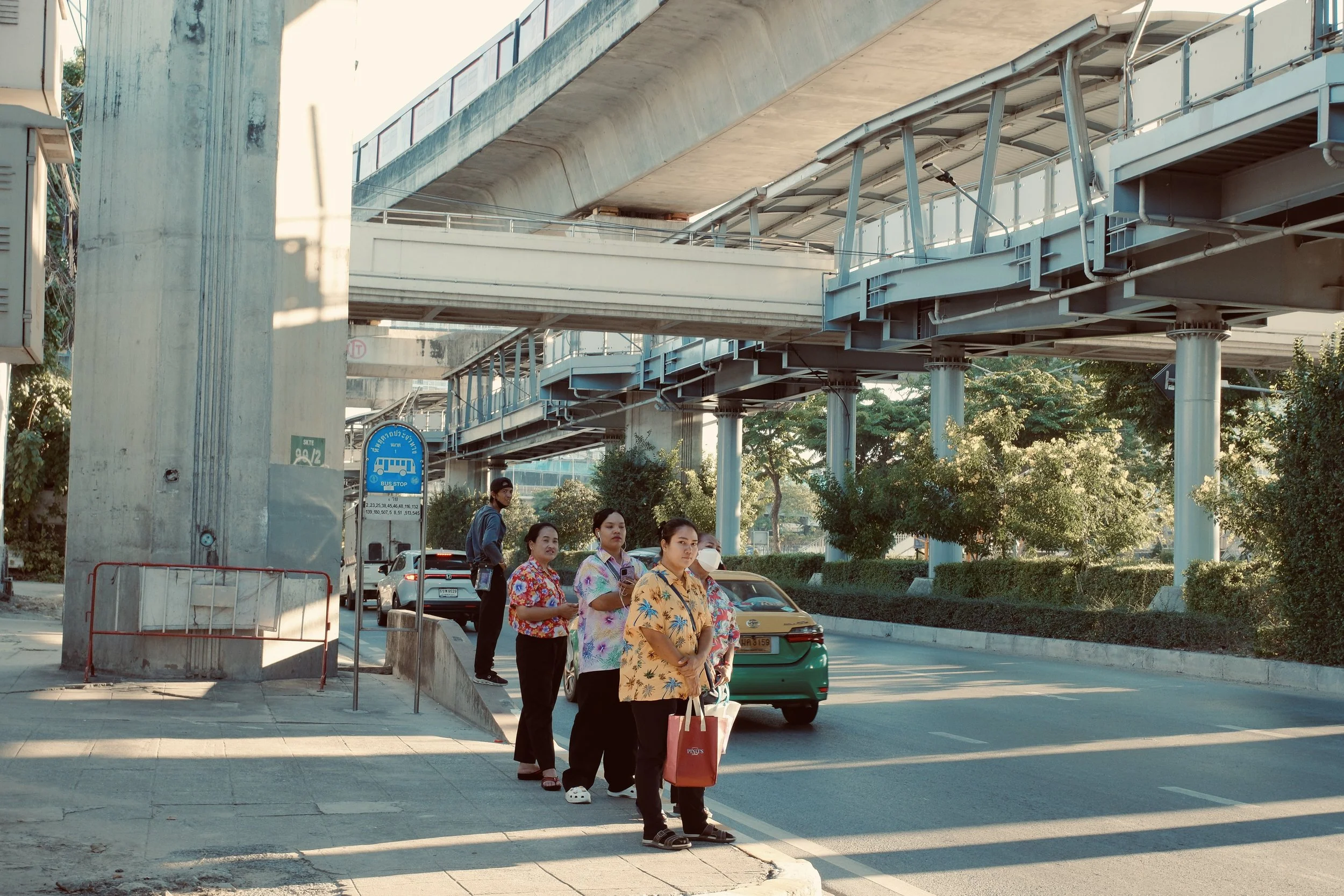 People wait in line at a bus stop under an elevated train track in an urban area during daytime.