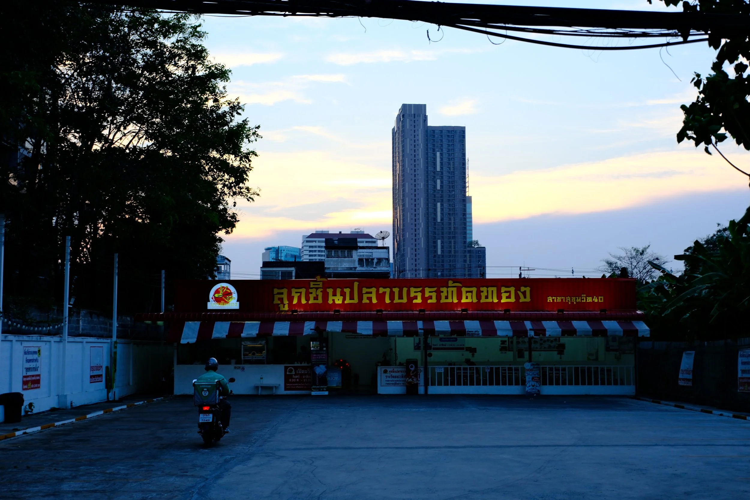 Empty parking lot outside a food stand with a red sign in Thai script, tall city buildings in the background, and trees framing the scene at sunset.