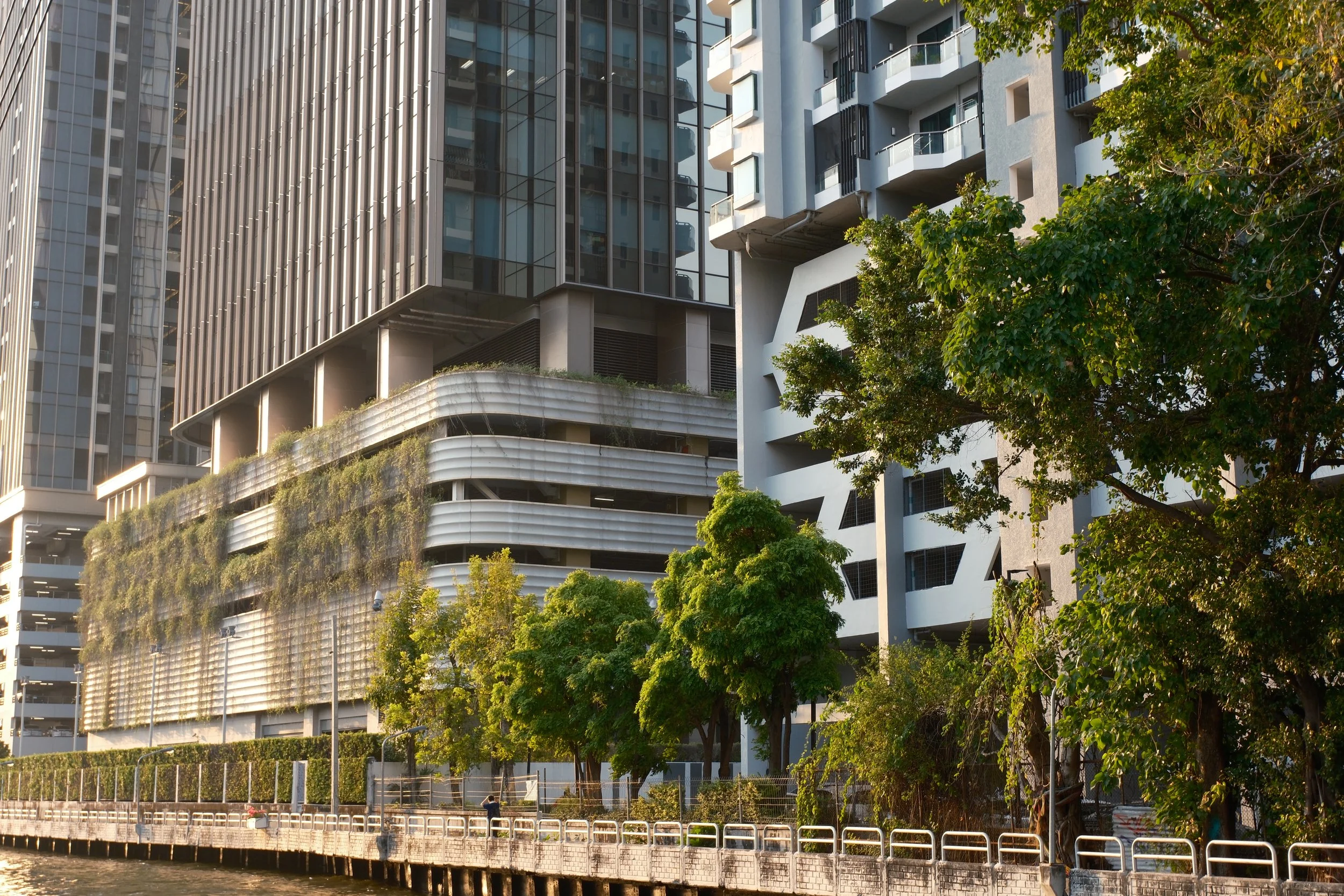 Modern high-rise buildings with glass and metal exteriors, trees along a riverbank, and a retaining wall in the foreground.