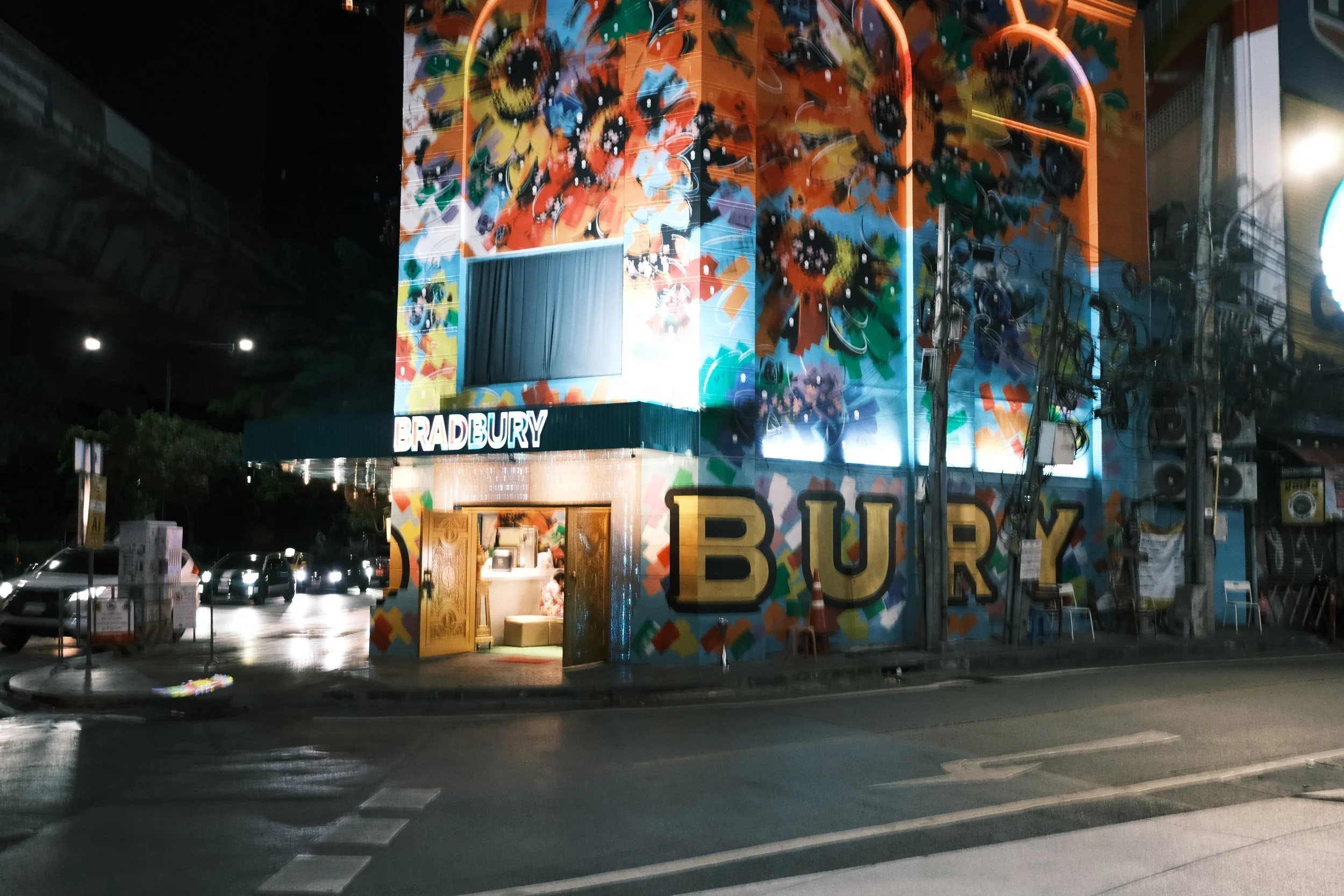 Night view of a building with colorful floral mural art, displaying the words 'BRADBURY' and large letters spelling 'BURY' on the facade. The building has a lit interior entrance and sits on a street with cars and some streetlights.