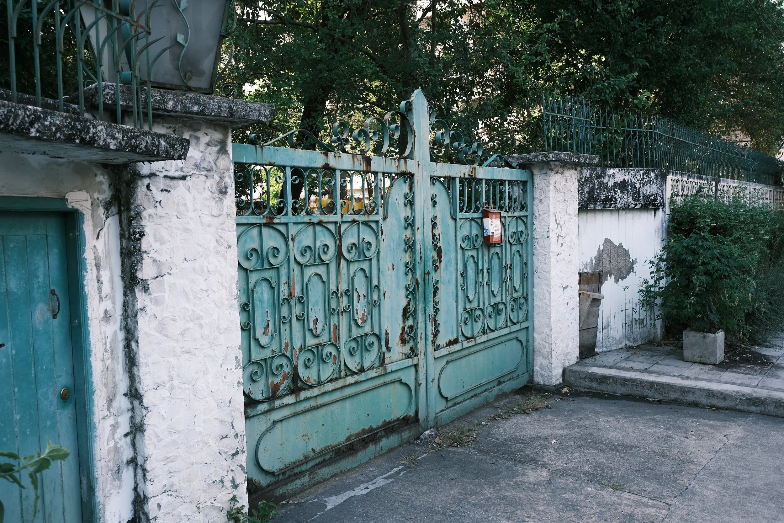 A weathered, turquoise wrought iron gate with intricate scrollwork, flanked by white stone pillars with moss and dirt, leading to a garden area with trees and bushes in the background.