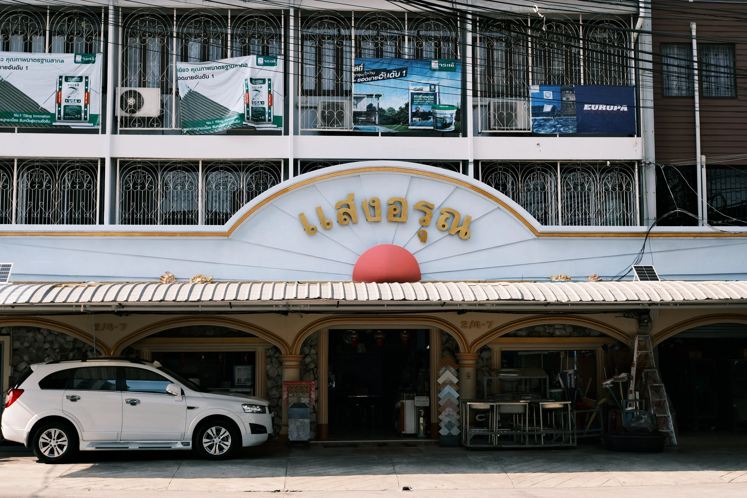 Front view of a building with a restaurant sign in Thai language, balconies with metal railings and advertisements, a white SUV parked in front, and some outdoor kitchen equipment.