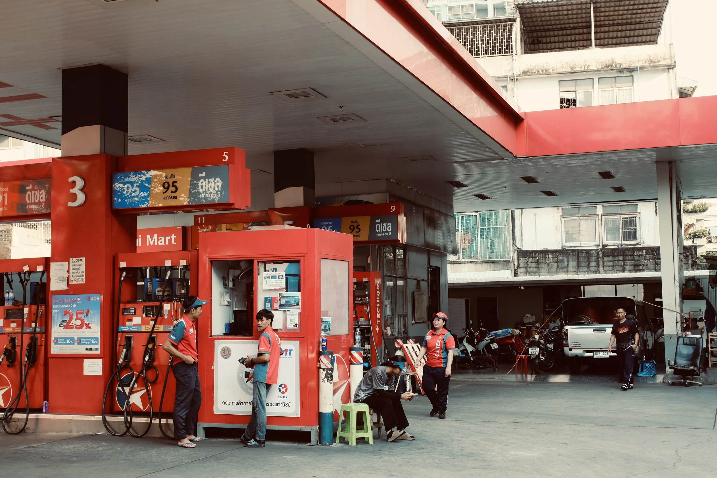 A gas station with three people wearing red shirts and dark pants, one sitting on a small green stool and two standing nearby, a white vehicle and motorcycles in the background, and a part of a building visible.