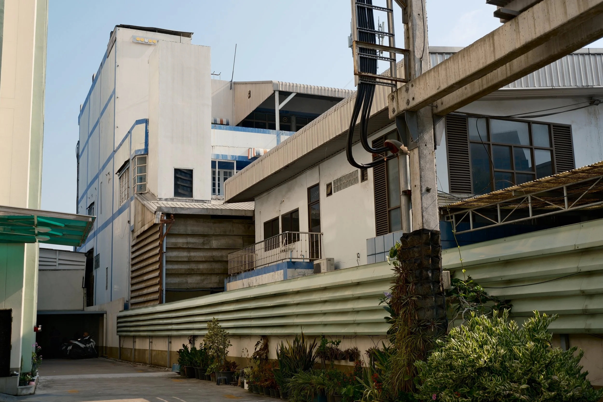 Several multi-story buildings with various windows, balconies, and exterior features in an urban setting, with plants along the ground and a parking area with a motorcycle.
