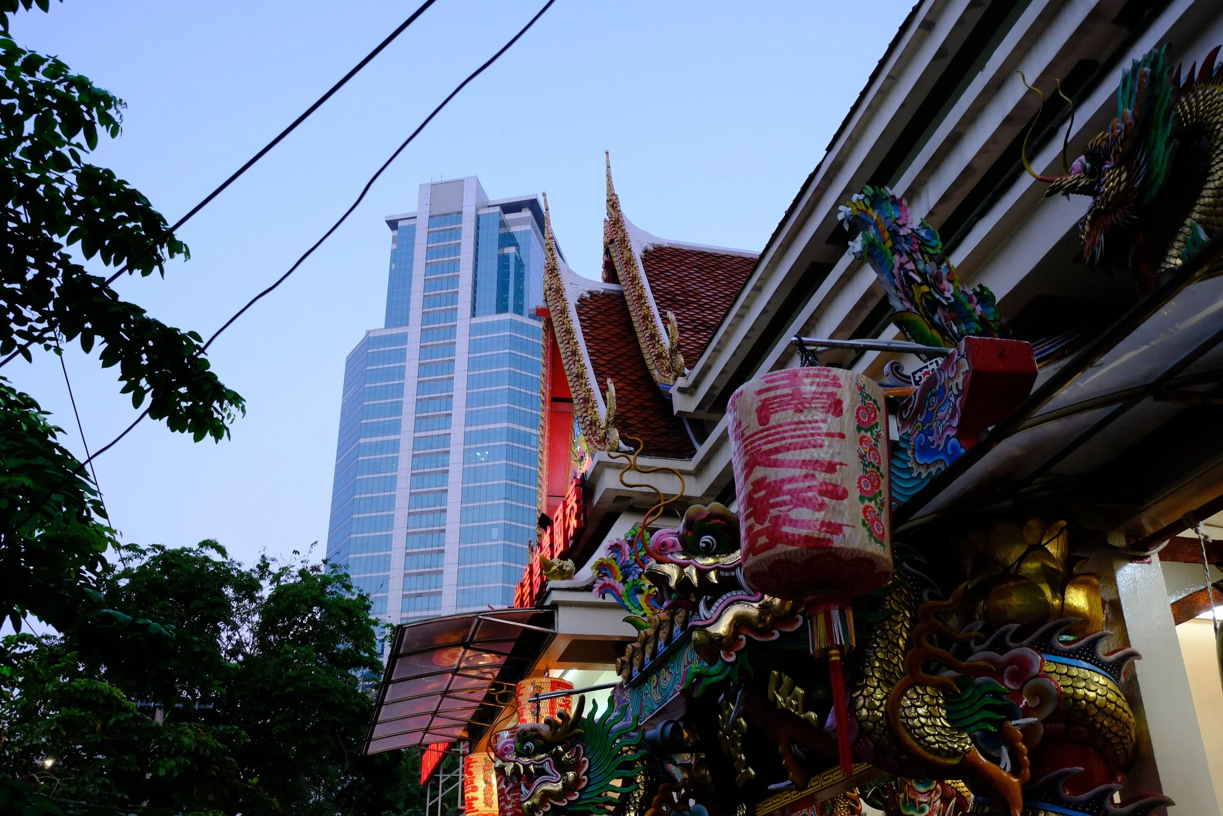 A traditional Thai temple with ornate dragon decorations, red lanterns, and a modern high-rise building in the background.