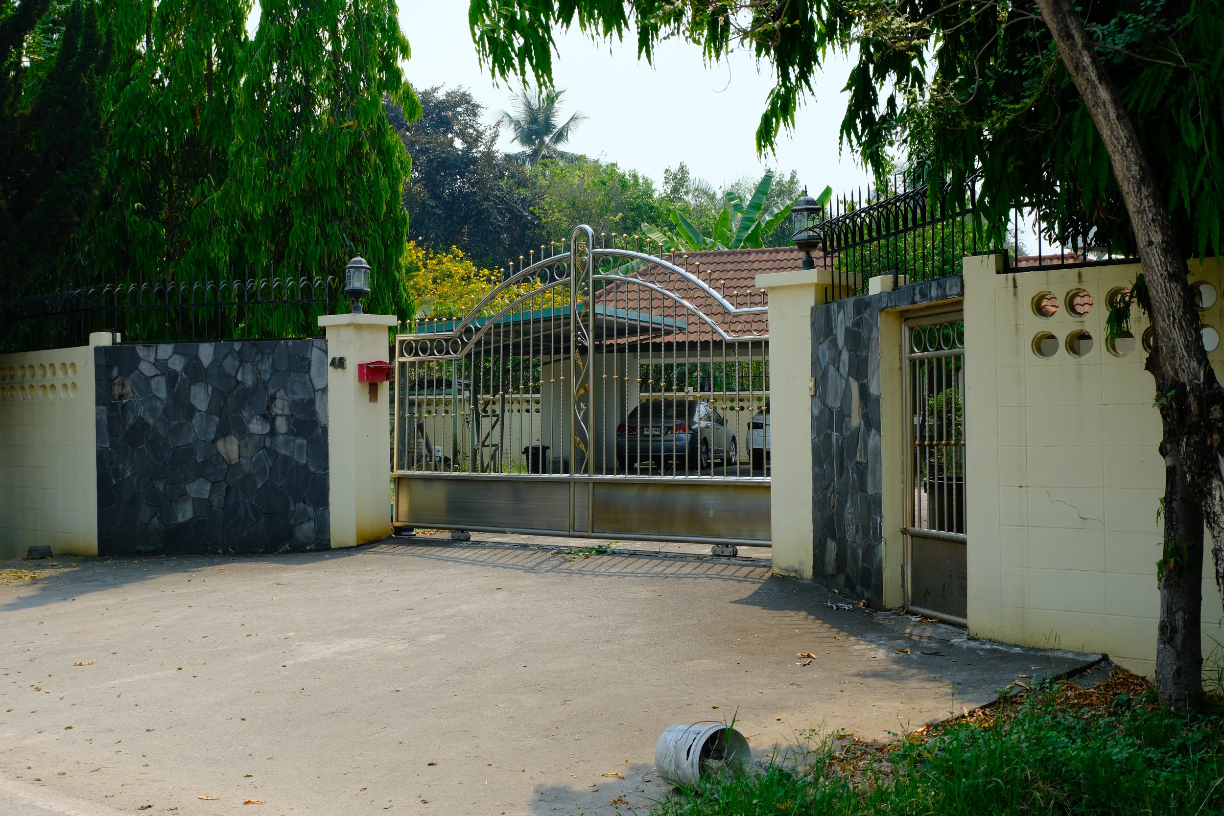 Gated entrance to a residential property with a metal gate, stone and concrete walls, and parked cars inside. Tall trees and lush greenery surround the gate.