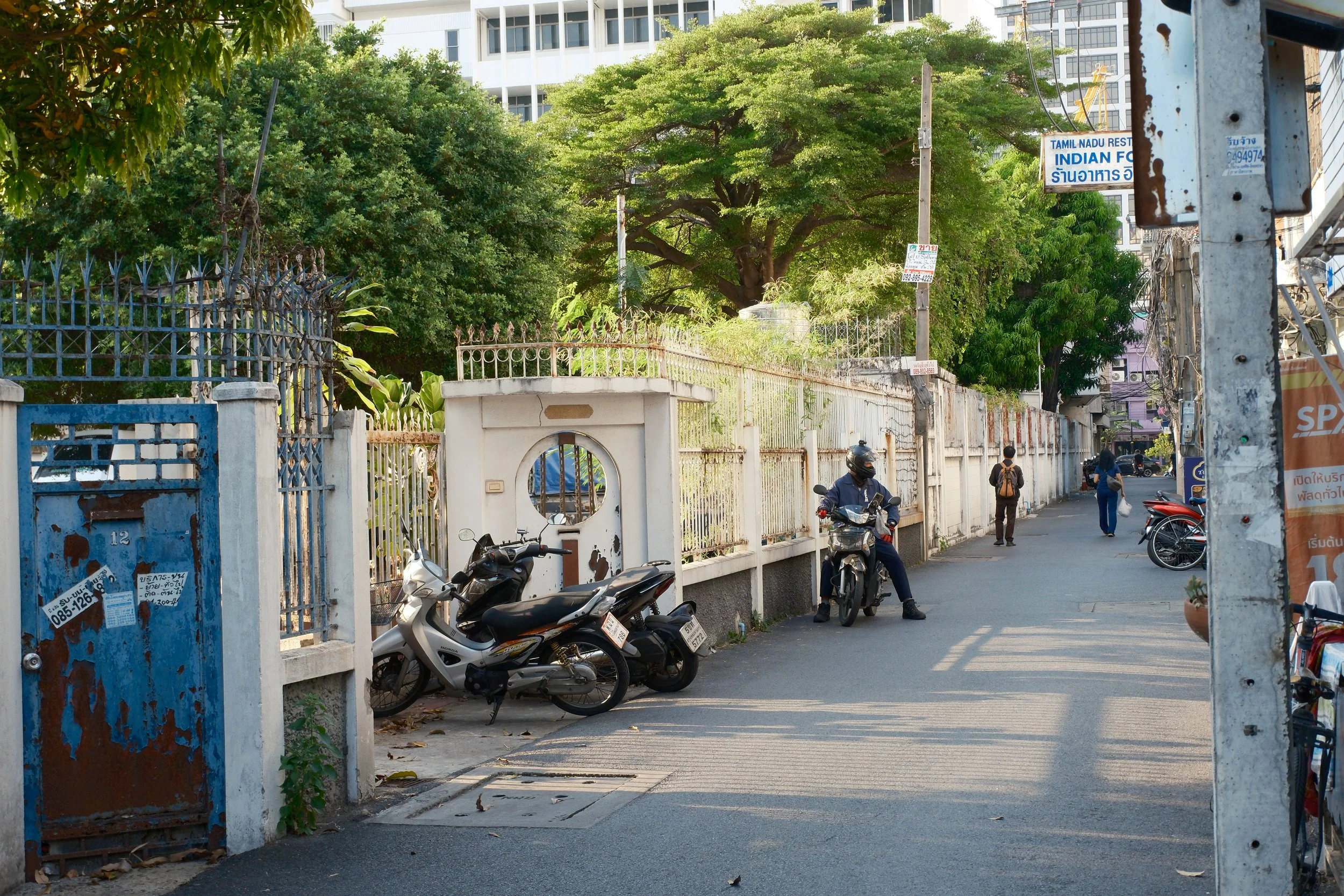 Urban street scene in Thailand with motorcycles parked near a white fence, a man on a scooter, and pedestrians walking, with trees and buildings in the background.