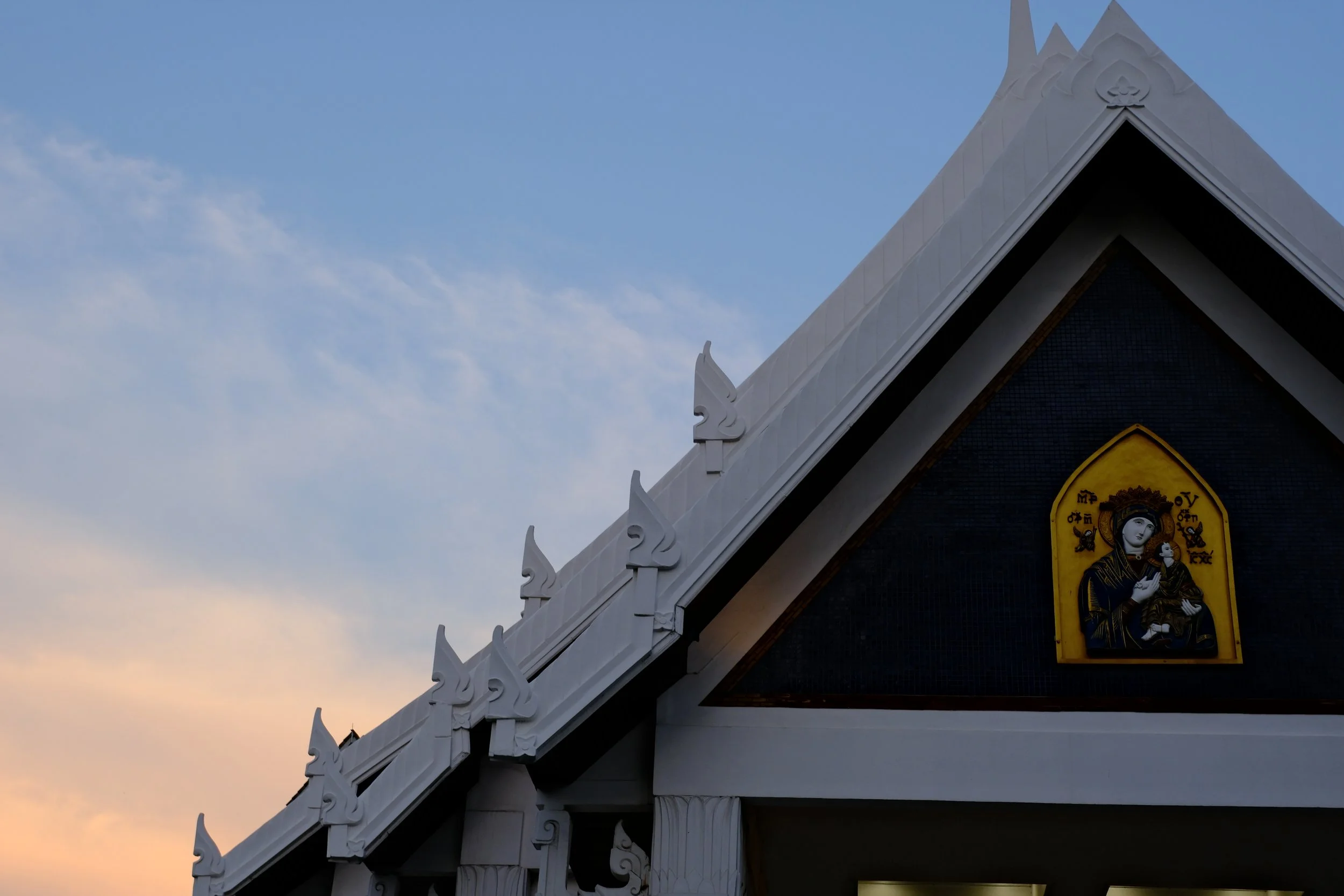 Close-up of the ornate roof and a religious icon on the gable of a temple or church, with a blue sky and light clouds in the background.