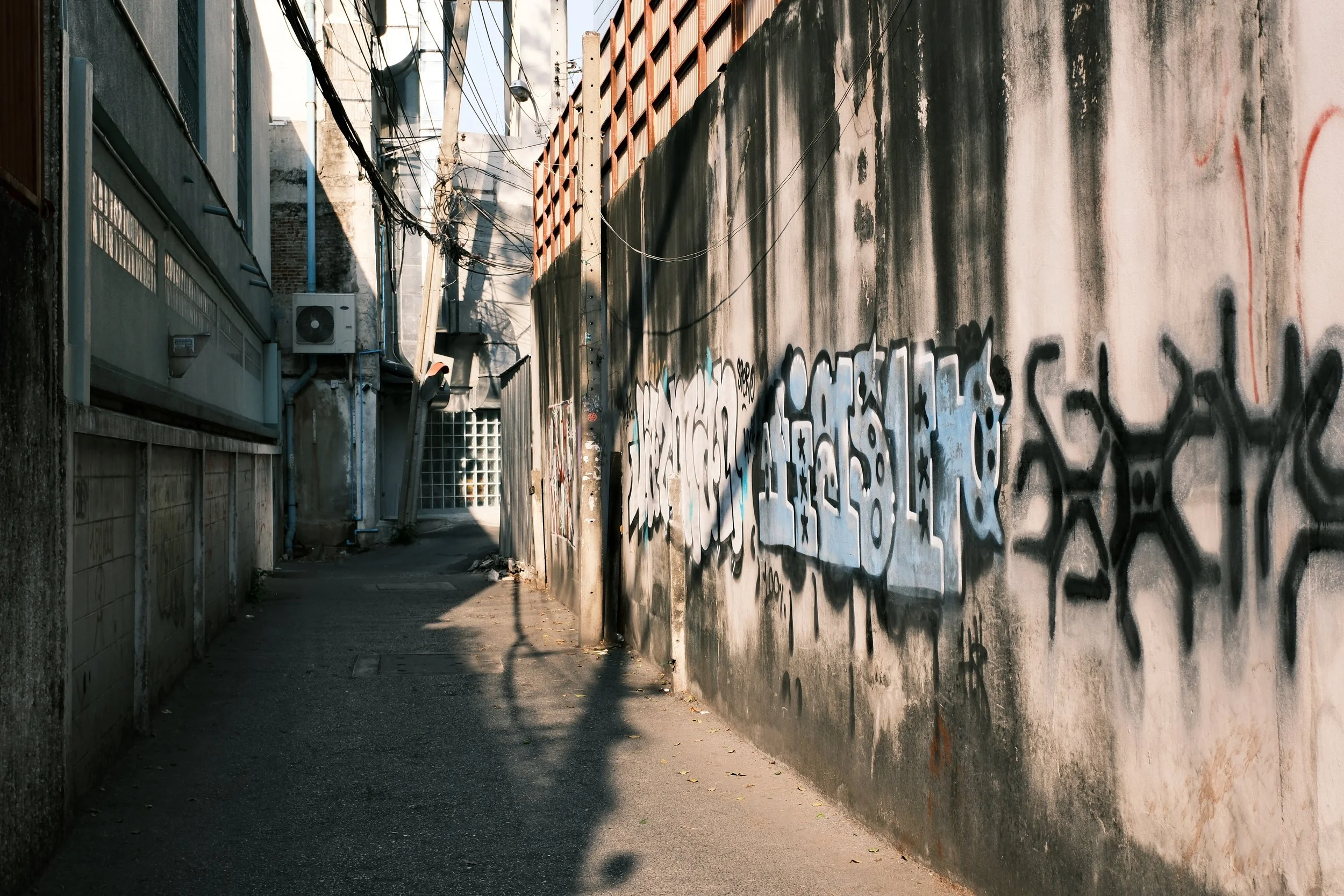 Alleyway with graffiti-covered wall, dirt ground, and utility wires overhead.
