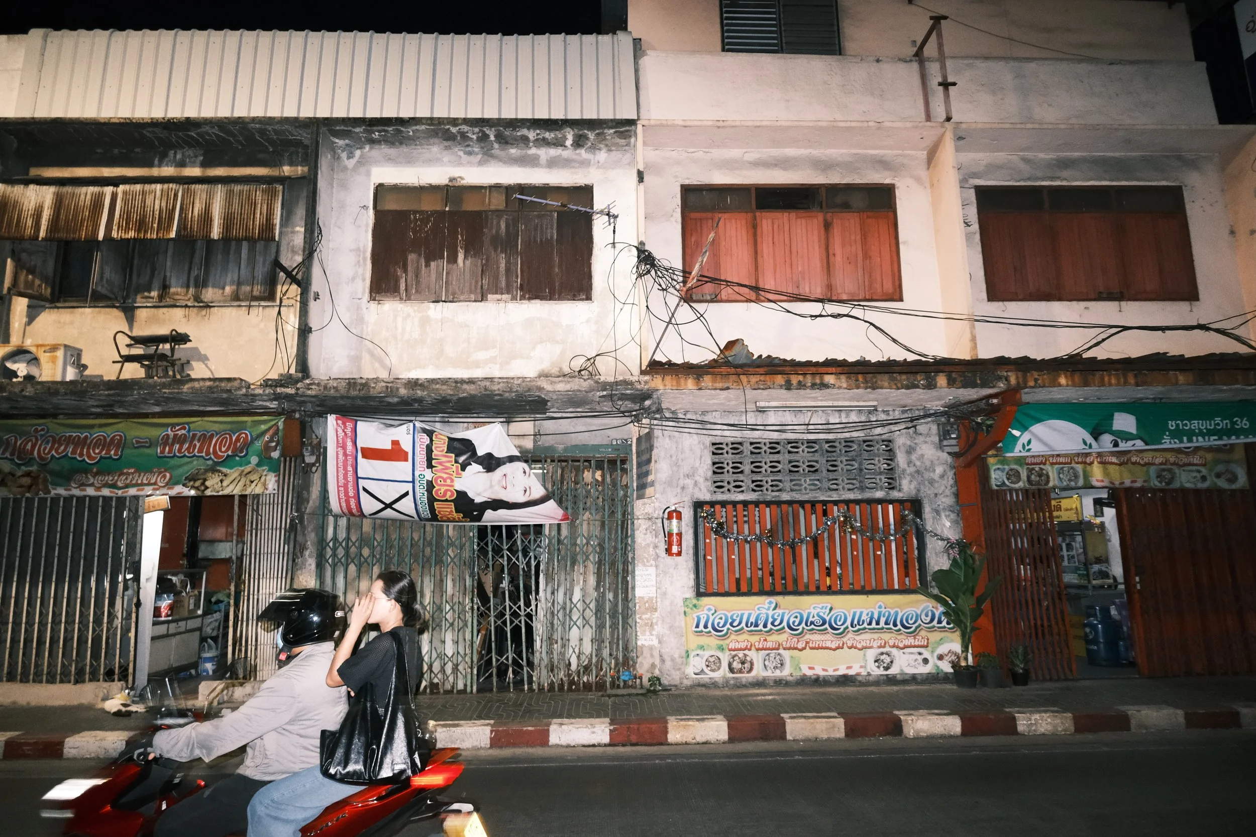 Night scene of old building facades with closed shutter windows, electrical wires, and advertising banners, a woman riding a scooter past the storefronts.