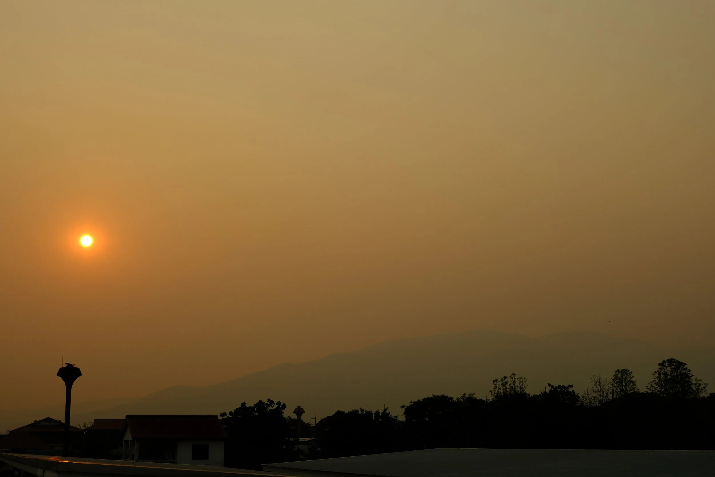 Hazy sunset or sunrise over a suburban area with silhouetted houses, trees, and hills in the distance. The sun is visible through a layer of haze or smog, casting a yellow-orange glow in the sky.