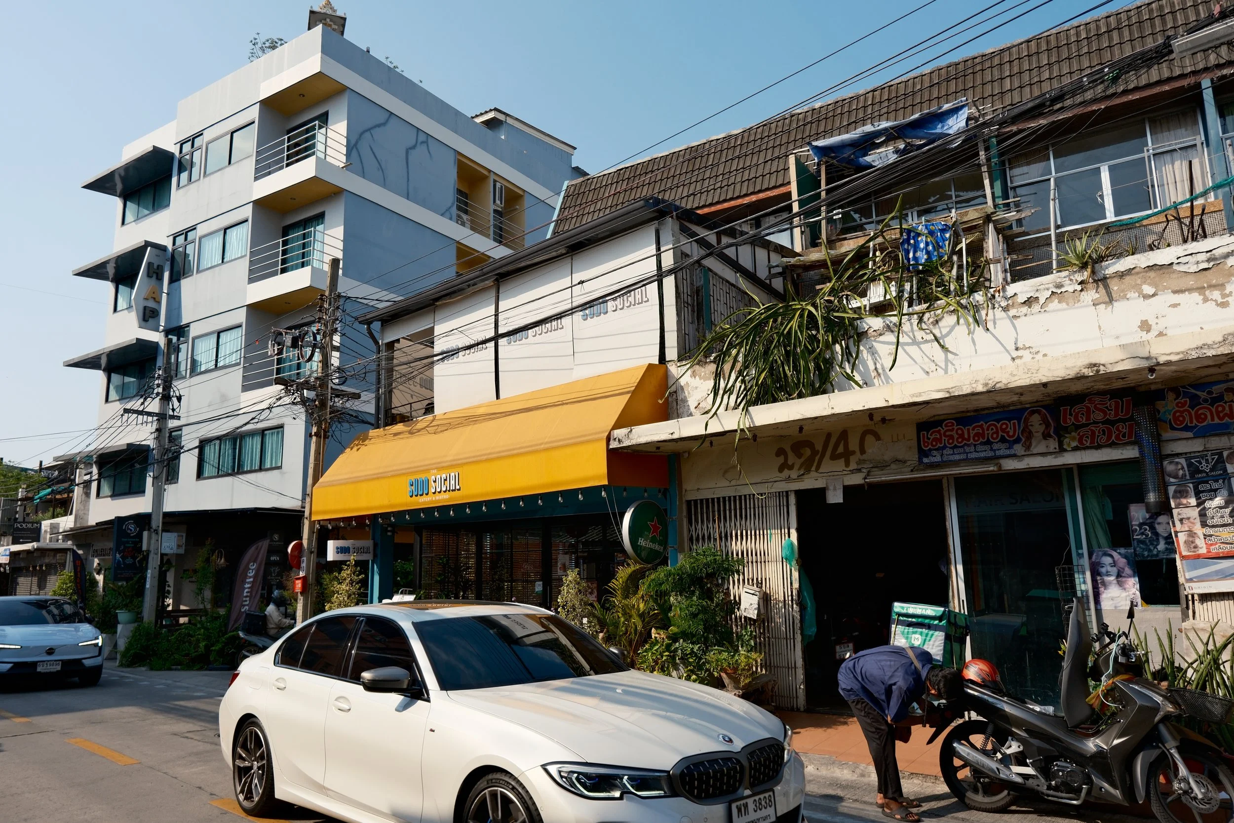 Street scene with parked cars, including a white BMW, a coffee-colored car, and a black scooter. Buildings with signage in Thai, a yellow awning, potted plants, and a person working on the scooter. Overhead power lines against a clear sky.