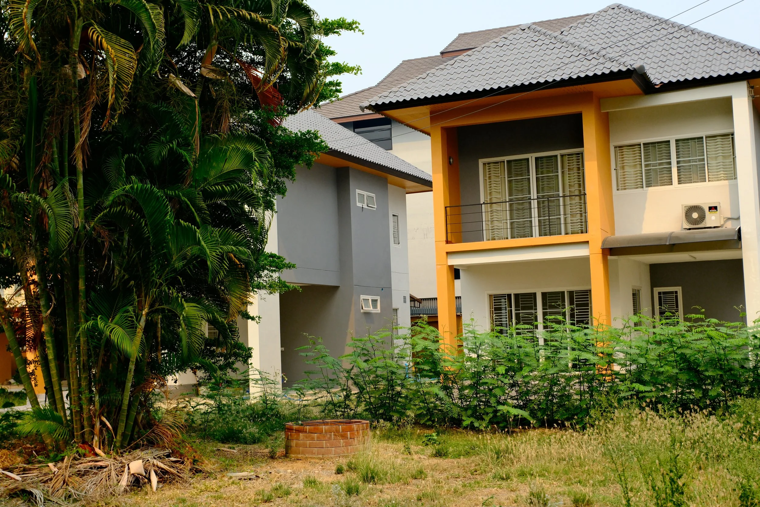 A modern two-story house with a yellow and white exterior, gray tiled roof, large windows, and balcony, surrounded by green bushes and a tree, with a grassy yard in the foreground.