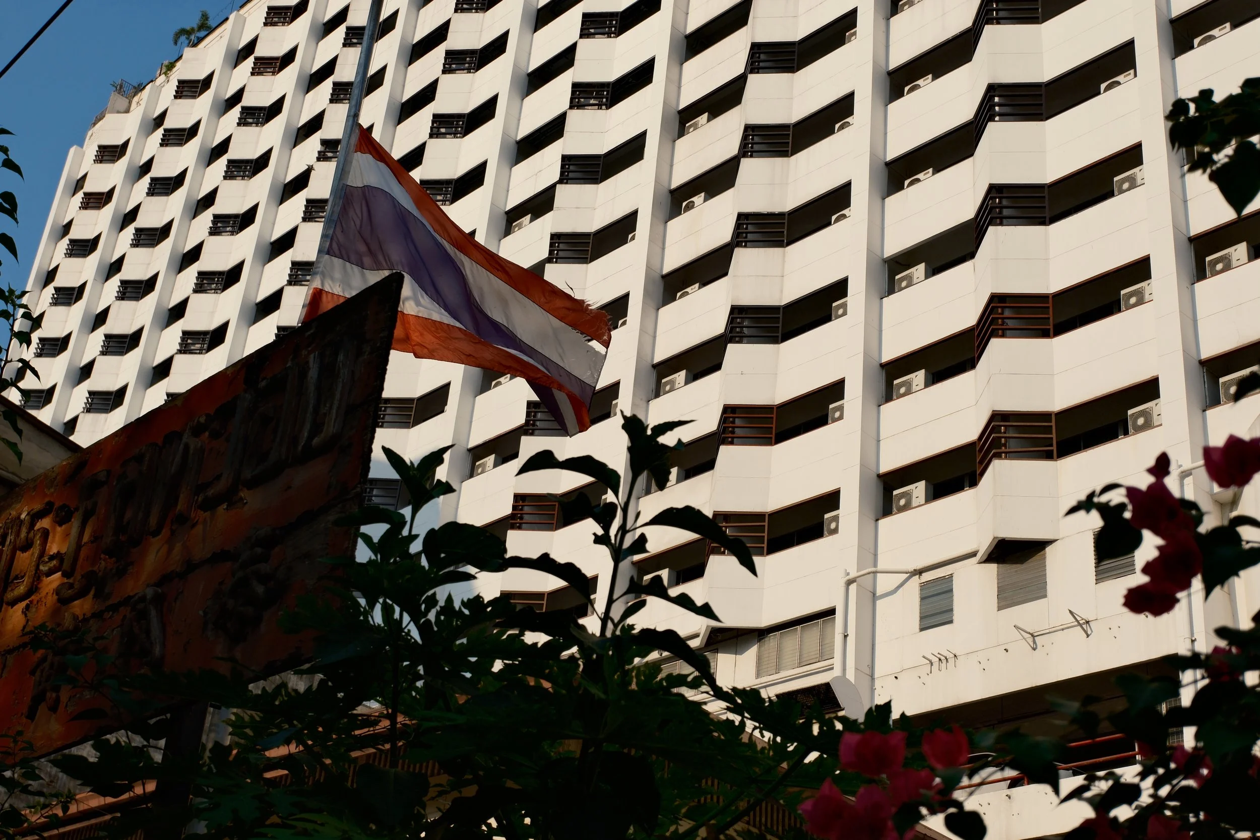 A tall white building with multiple small balconies and air conditioning units visible on the facade. A Thai flag is in the foreground, alongside some greenery and flowers.