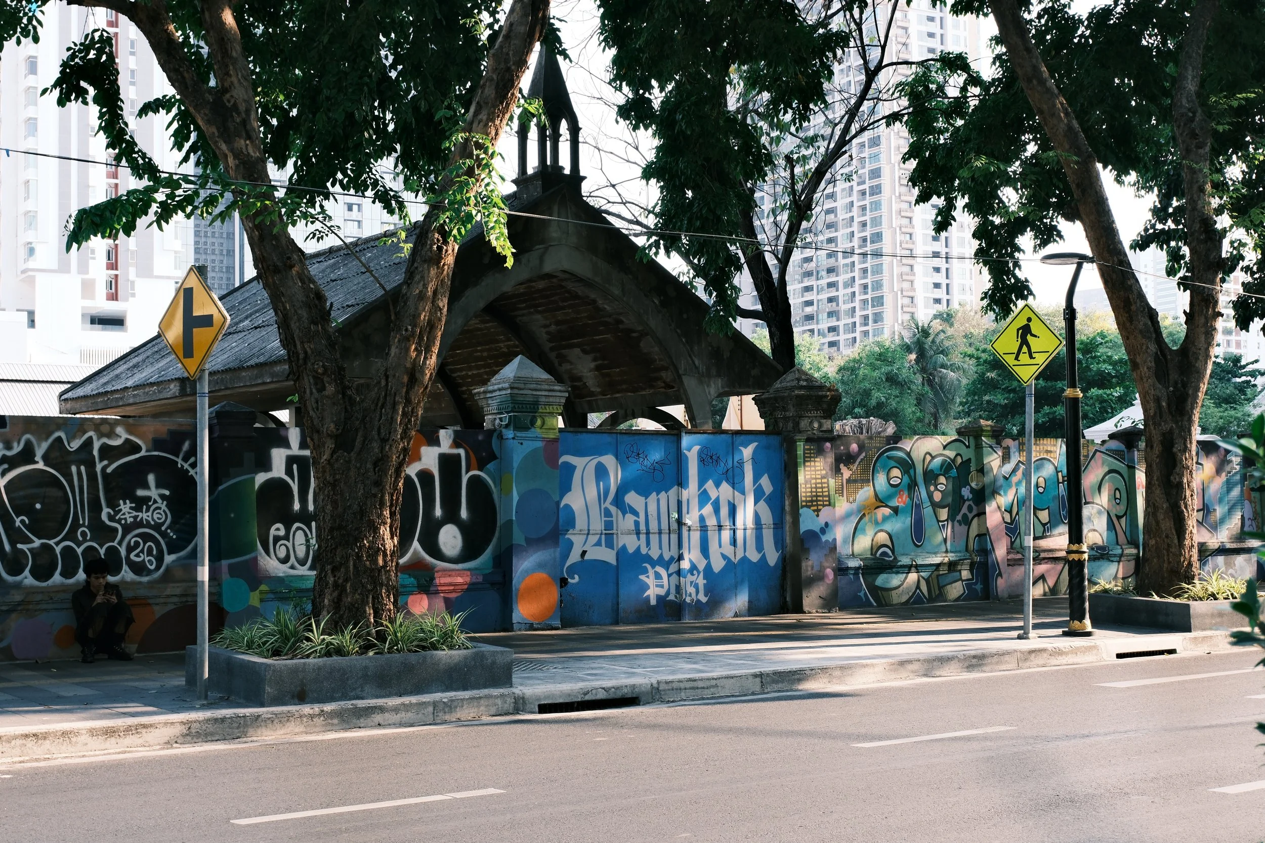 A street scene featuring a graffiti-covered wall behind two trees and modern high-rise buildings in the background. There are yellow pedestrian crossing signs and a person sitting on the sidewalk.