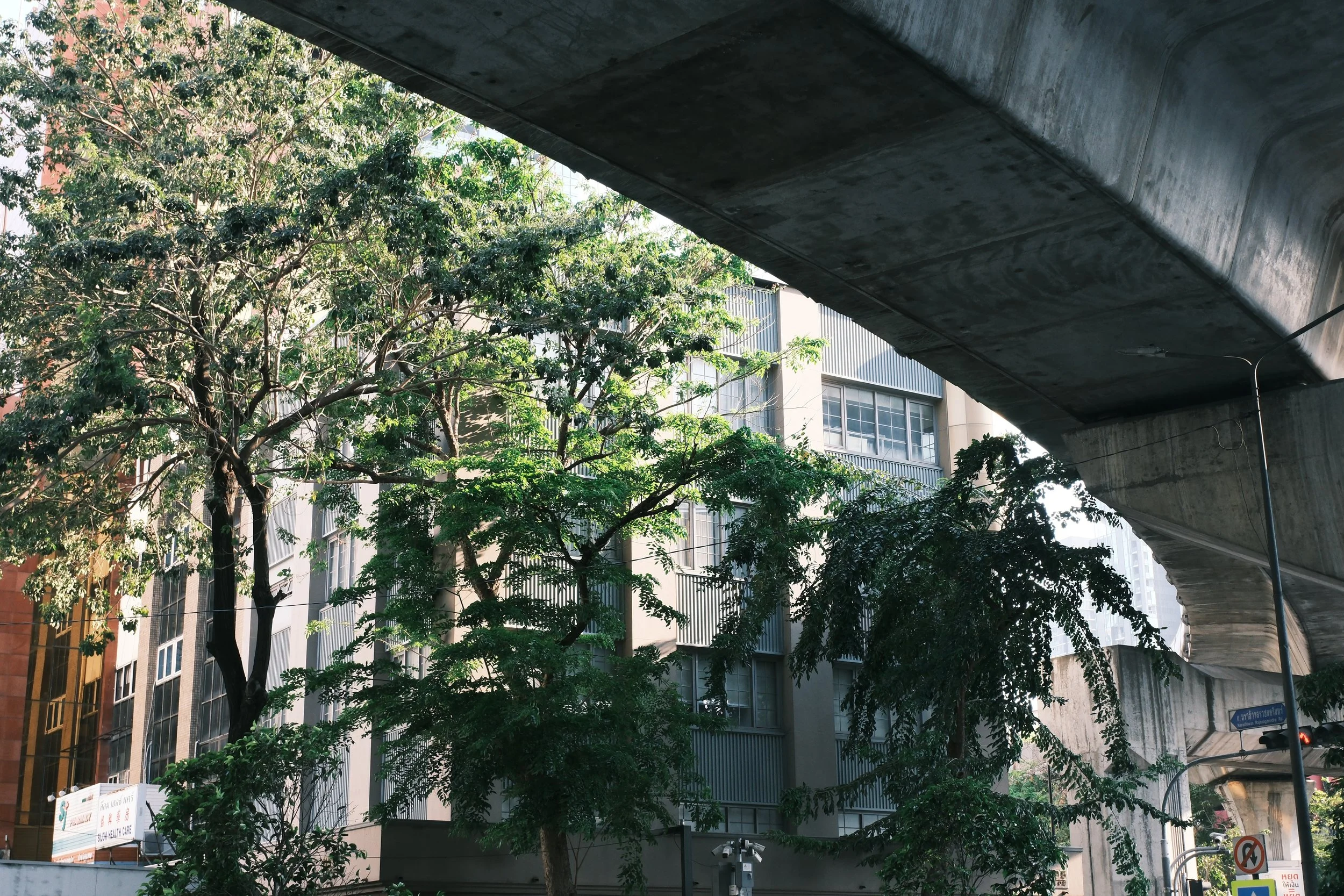 Urban scene with tall buildings, green trees, and an elevated concrete structure overhead.