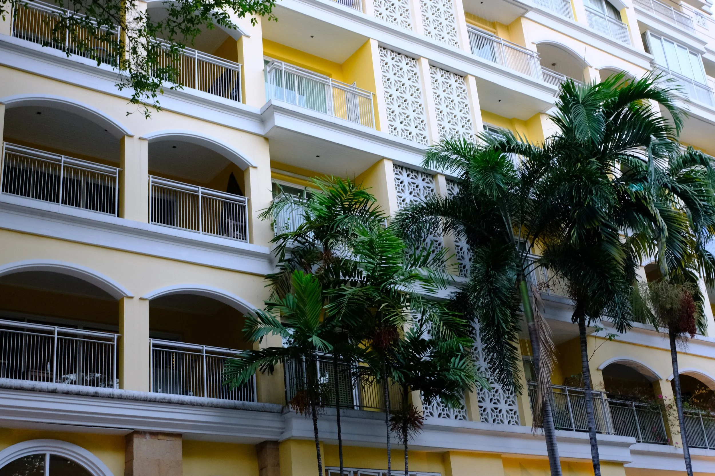 A multi-story yellow building with white decorative lattice panels on the sides, multiple balconies with white railings, and tropical palm trees in front.
