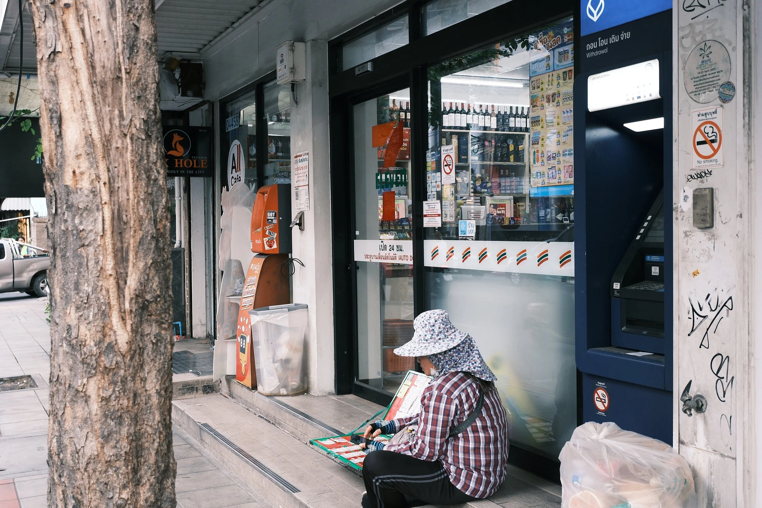 A person sitting on the sidewalk outside a convenience store, wearing a patterned hat and plaid jacket, with a large plastic bag beside them.