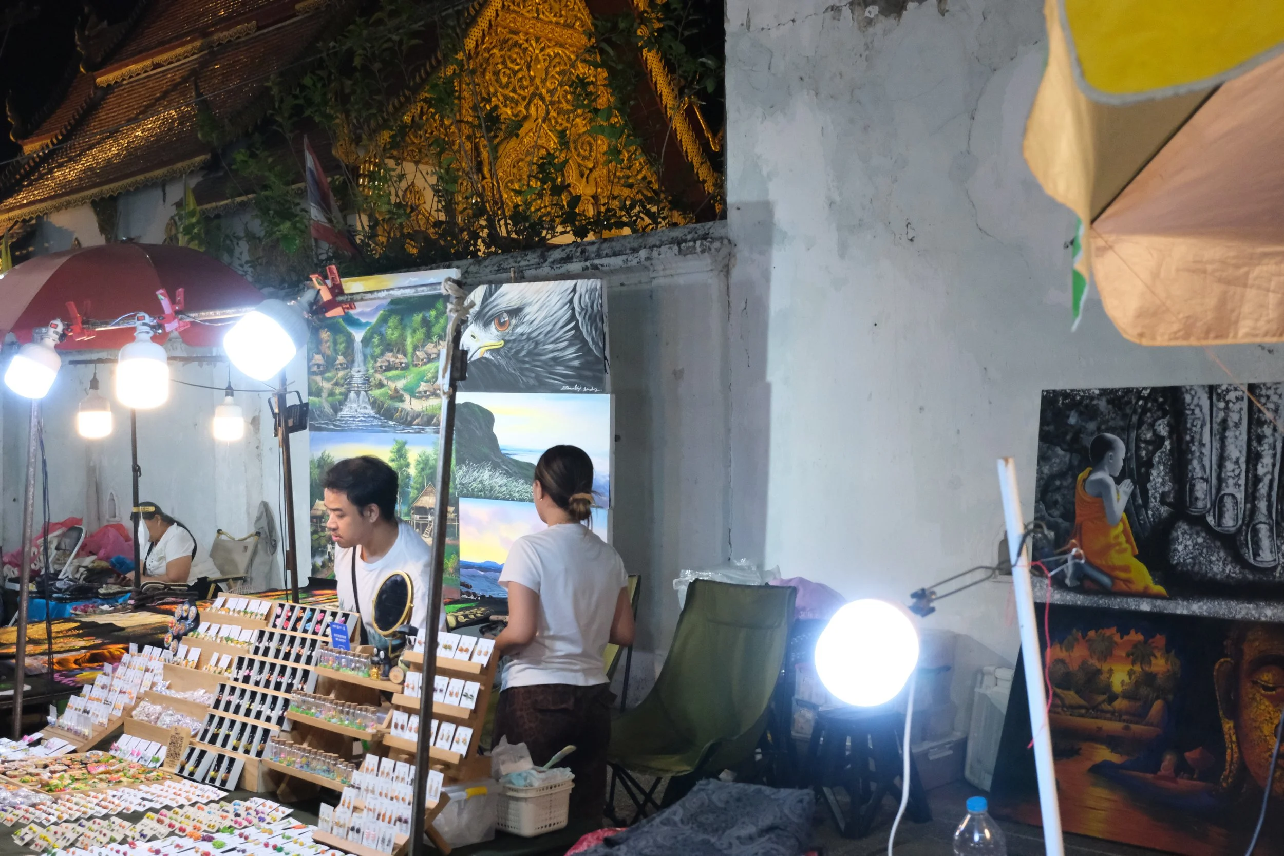 An outdoor art and craft market at night, with vendors displaying paintings and jewelry, illuminated by bright lights, with a few people browsing.