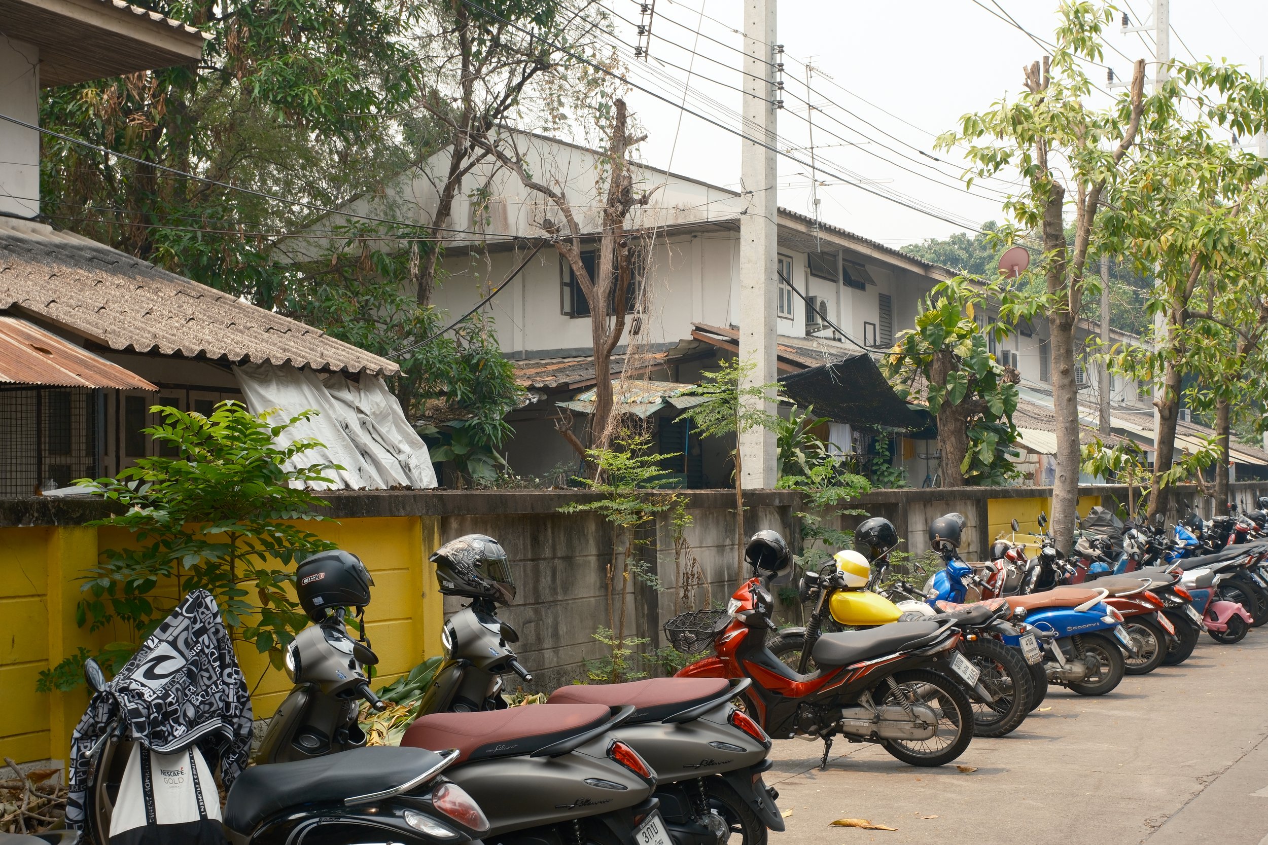 A row of parked motorcycles and scooters along a street in front of a concrete wall with houses and trees in the background.