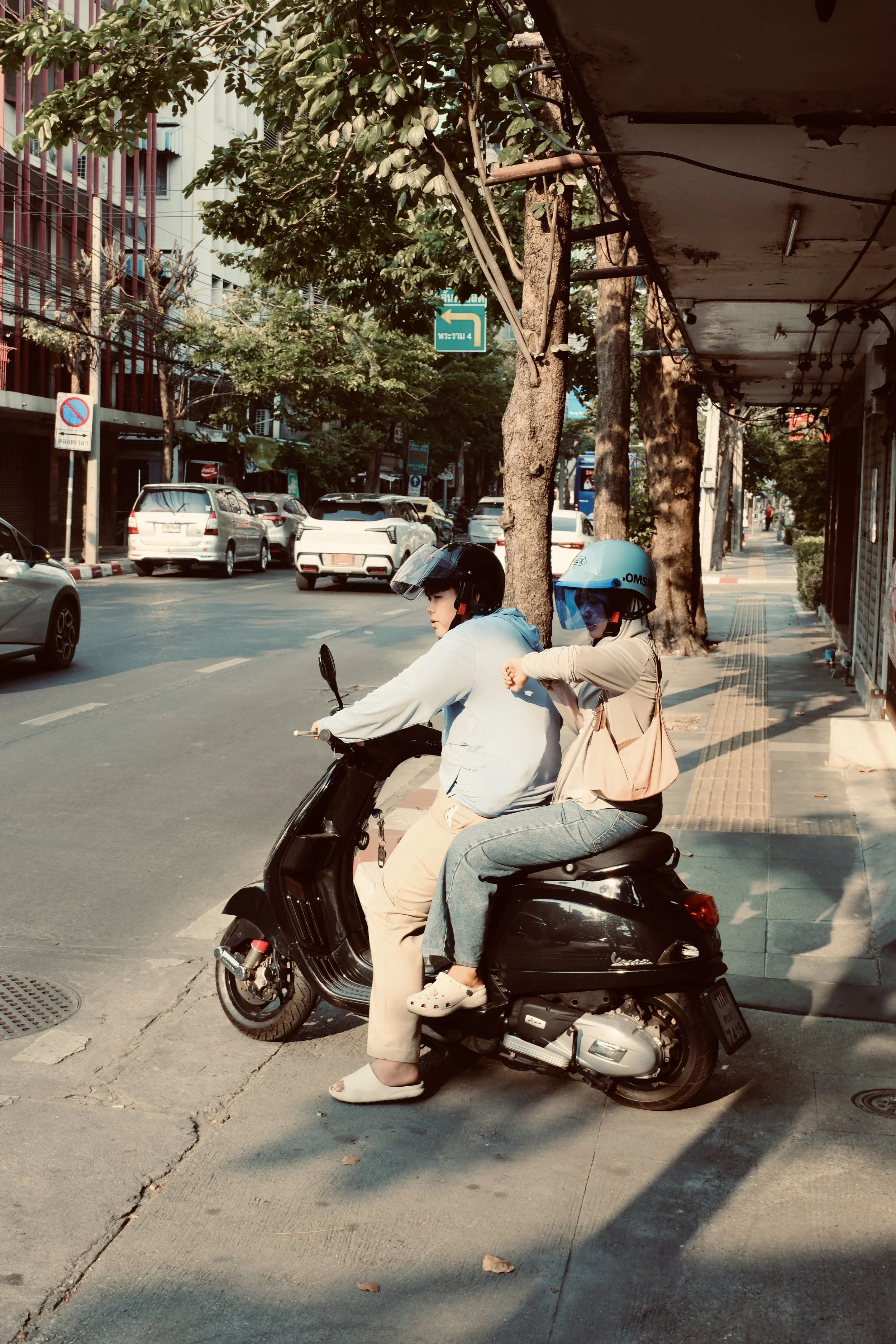 People riding a scooter on a city sidewalk, with one woman sitting behind another riding a scooter, both wearing helmets, in an urban street scene.