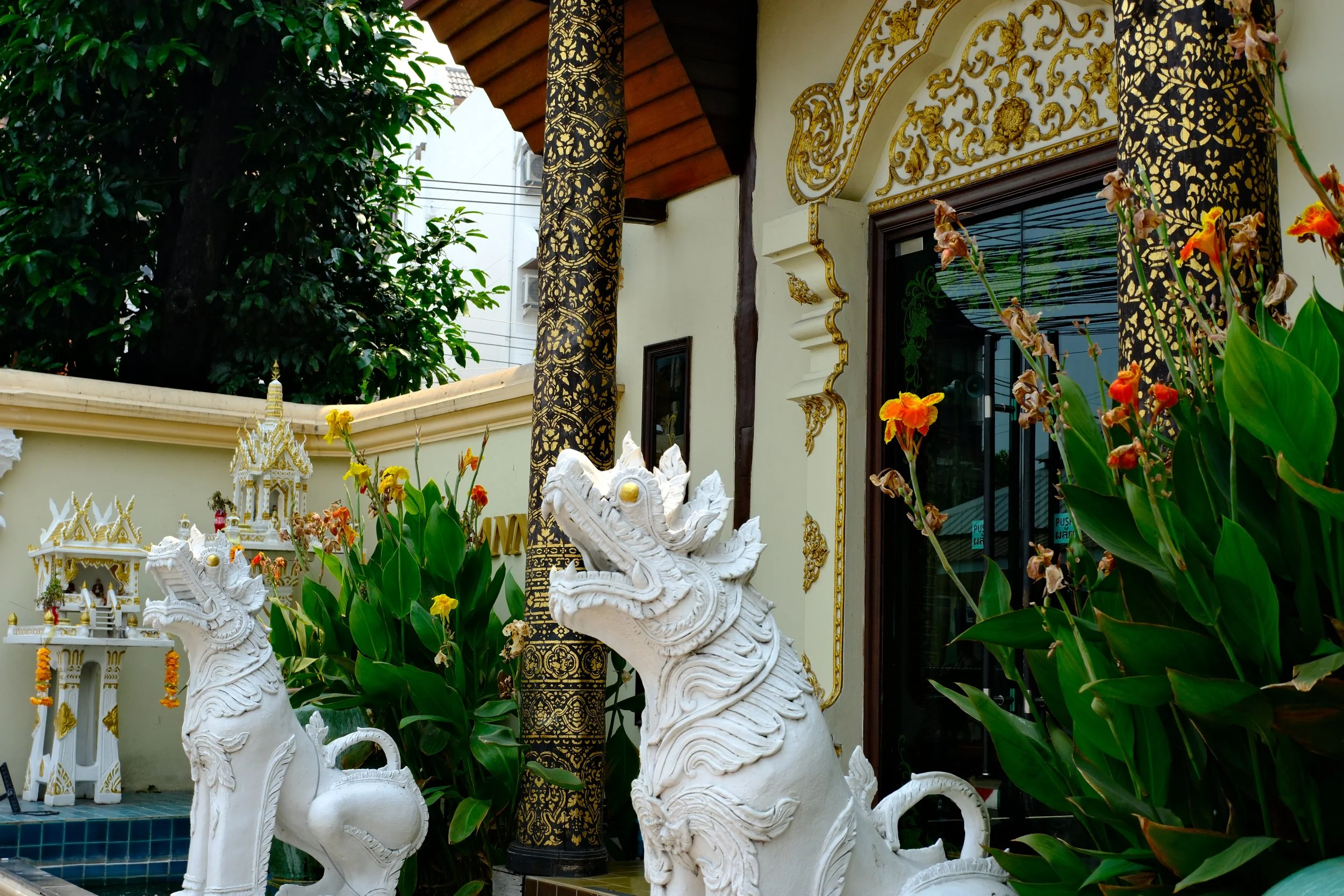 Decorative white statues of mythical creatures at the entrance of a building with ornate yellow and gold accents, surrounded by orange and yellow flowers and lush green plants.
