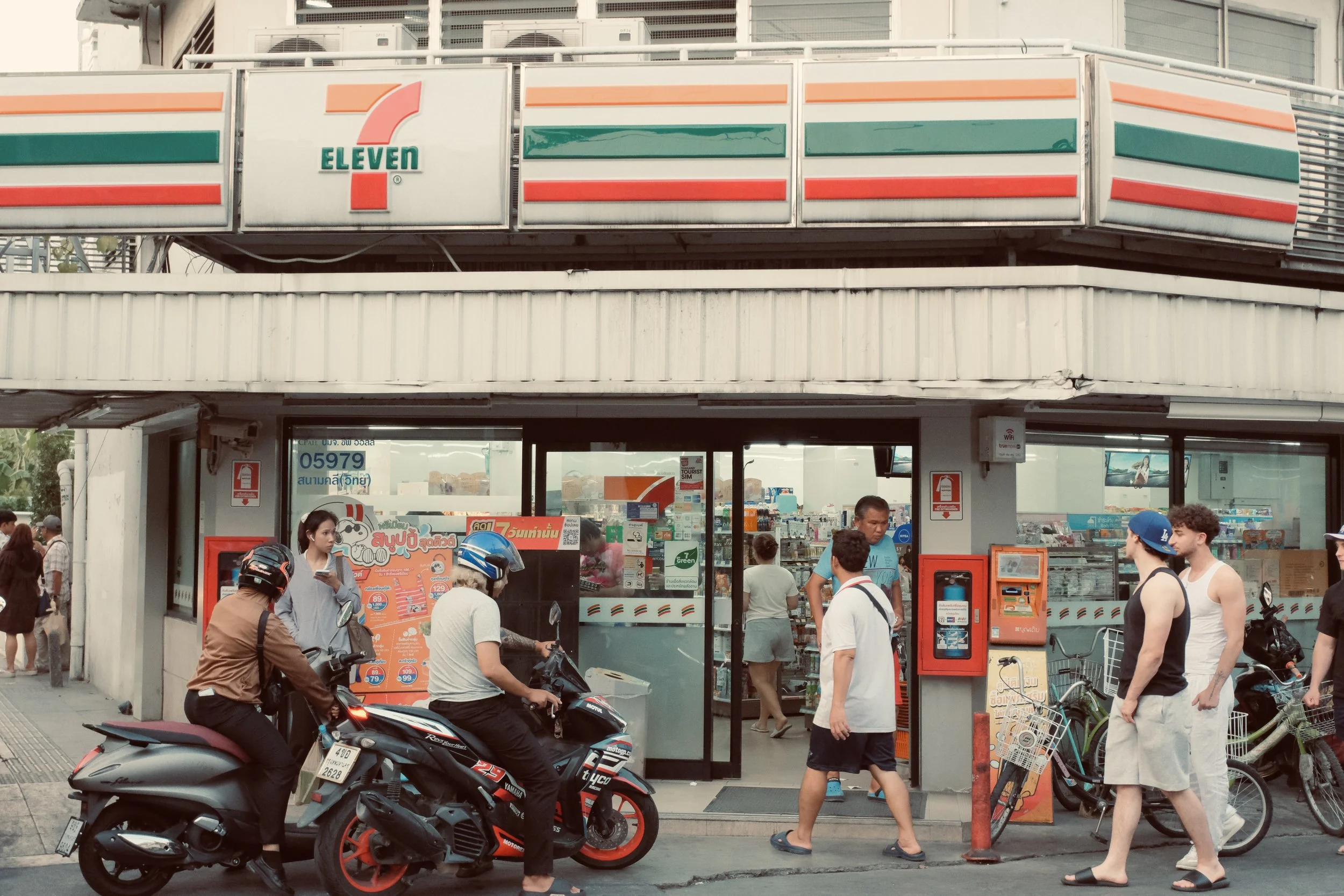 A 7-Eleven convenience store with people and motorbikes outside.