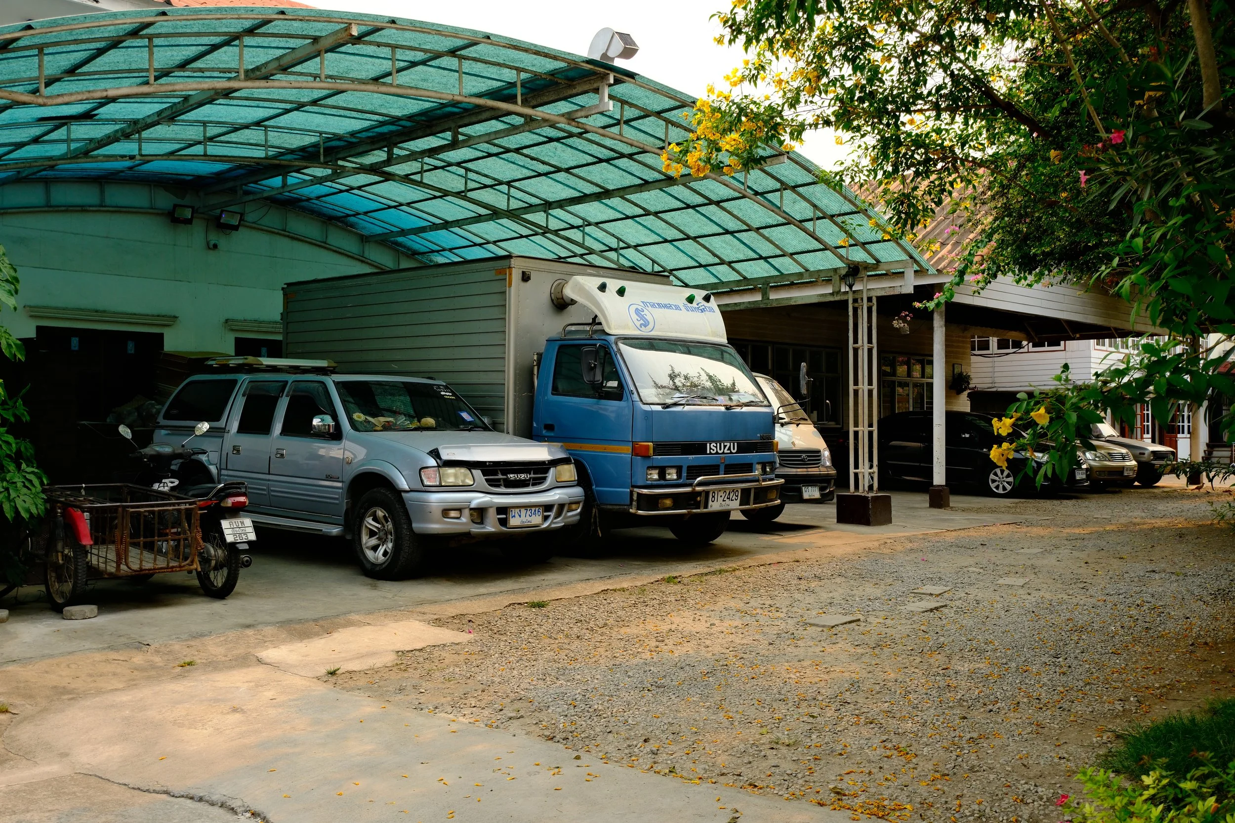 Various parked vehicles, including an Isuzu truck, an SUV, and a motorcycle, under a covered parking area with trees and flowers nearby.