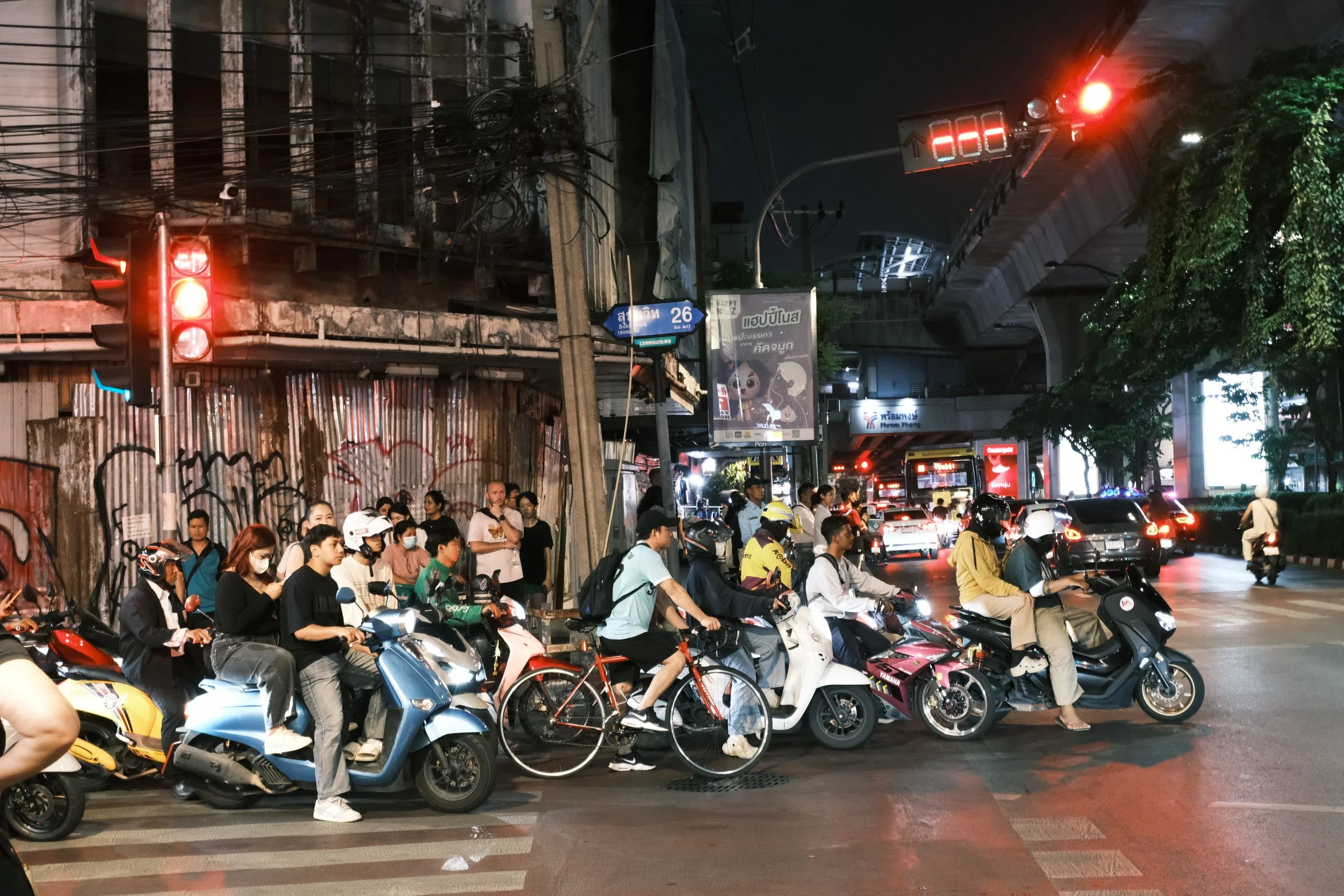 Nighttime city street scene with a crowd of people waiting to cross at a crosswalk, some on scooters and motorcycles, and others on bicycles or standing. Traffic lights are red, and there are vehicles on the road, including a bus and cars. The scene 