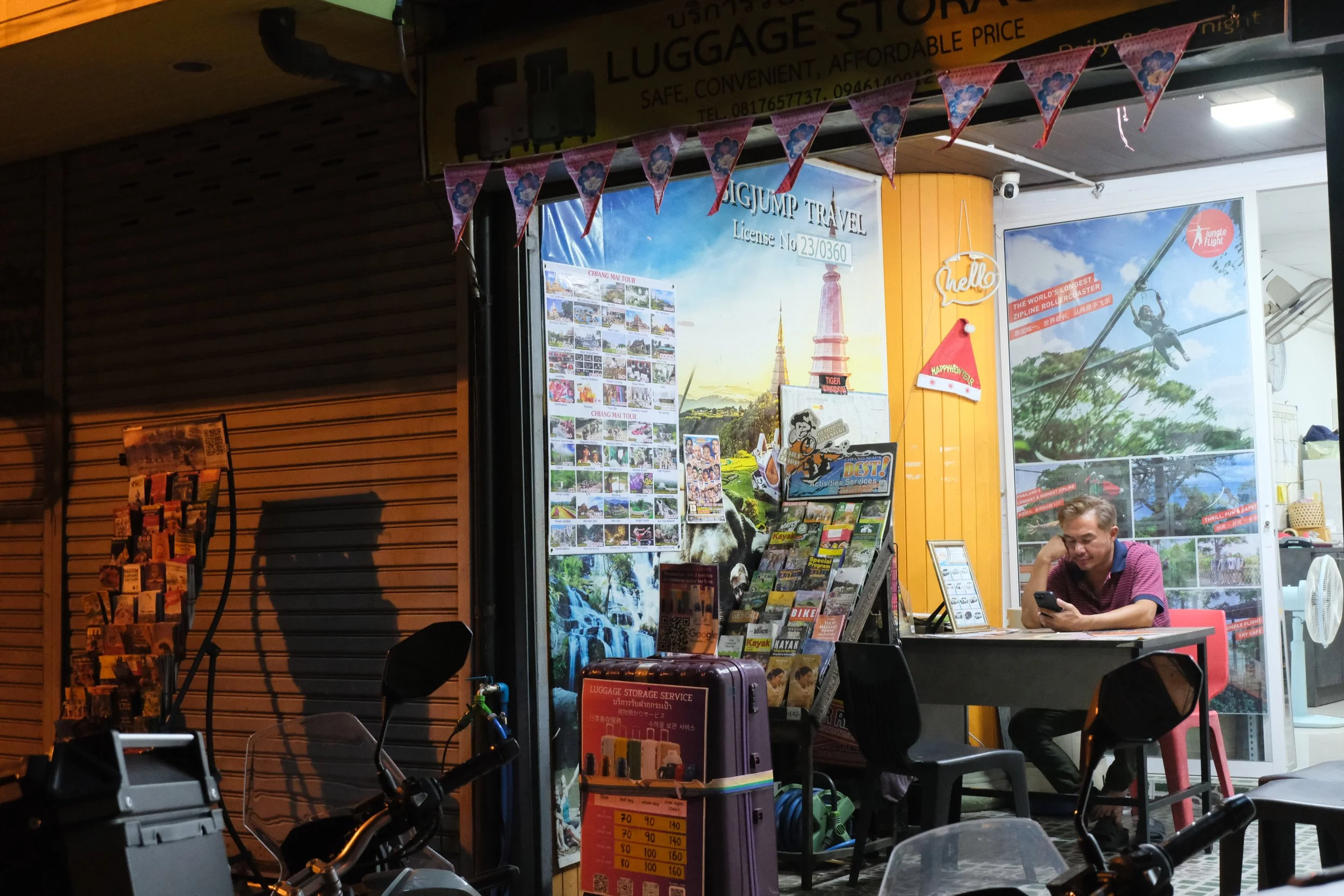 A man sitting at a table inside a travel agency, looking at his phone. The travel agency has colorful posters and brochures on the walls, and large posters advertising adventure activities like ziplining. There is a purple luggage storage container i