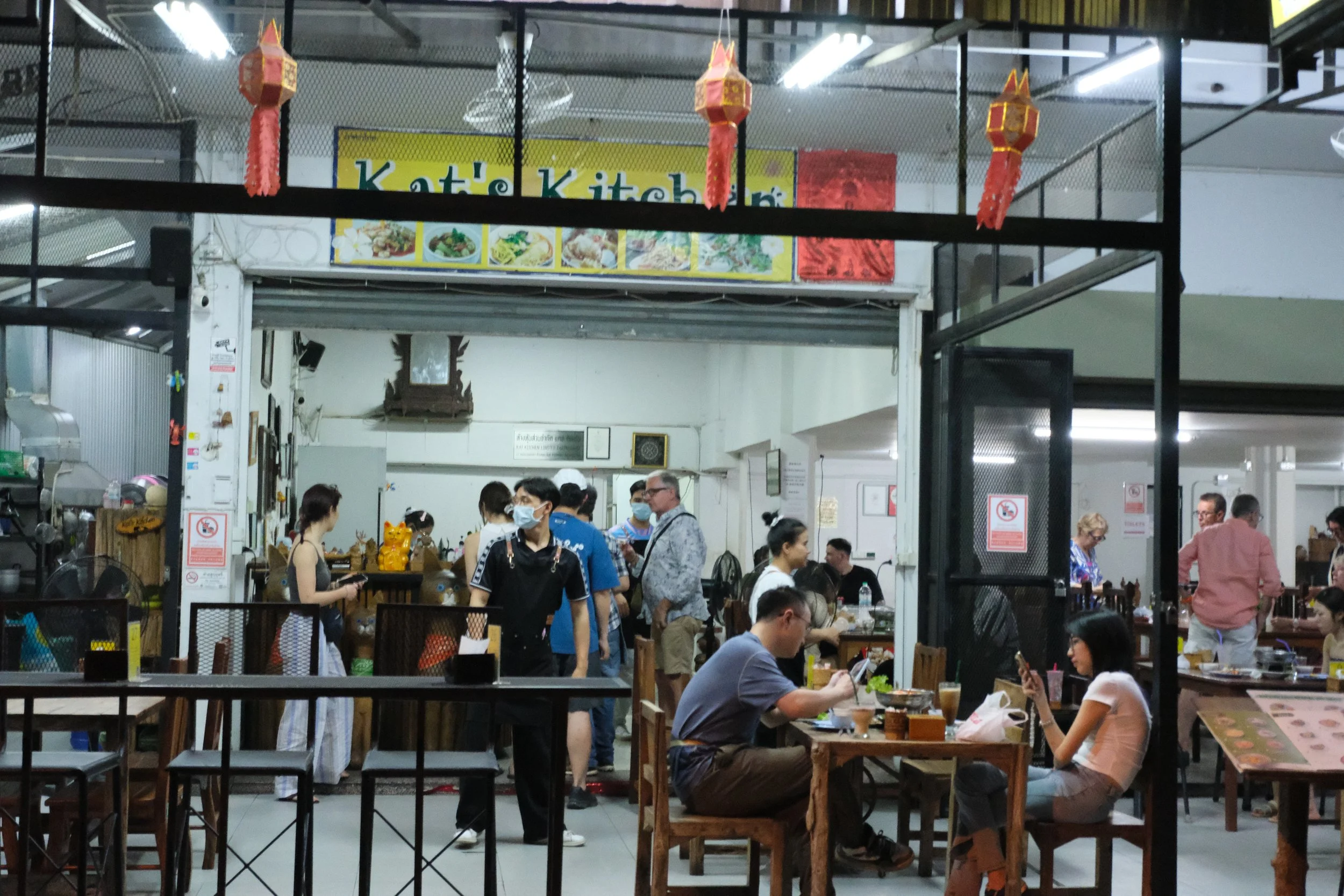 A busy indoor Asian restaurant with customers dining and staff working, decorated with red Chinese lanterns and a yellow sign reading 'Kati's Kitchen.'