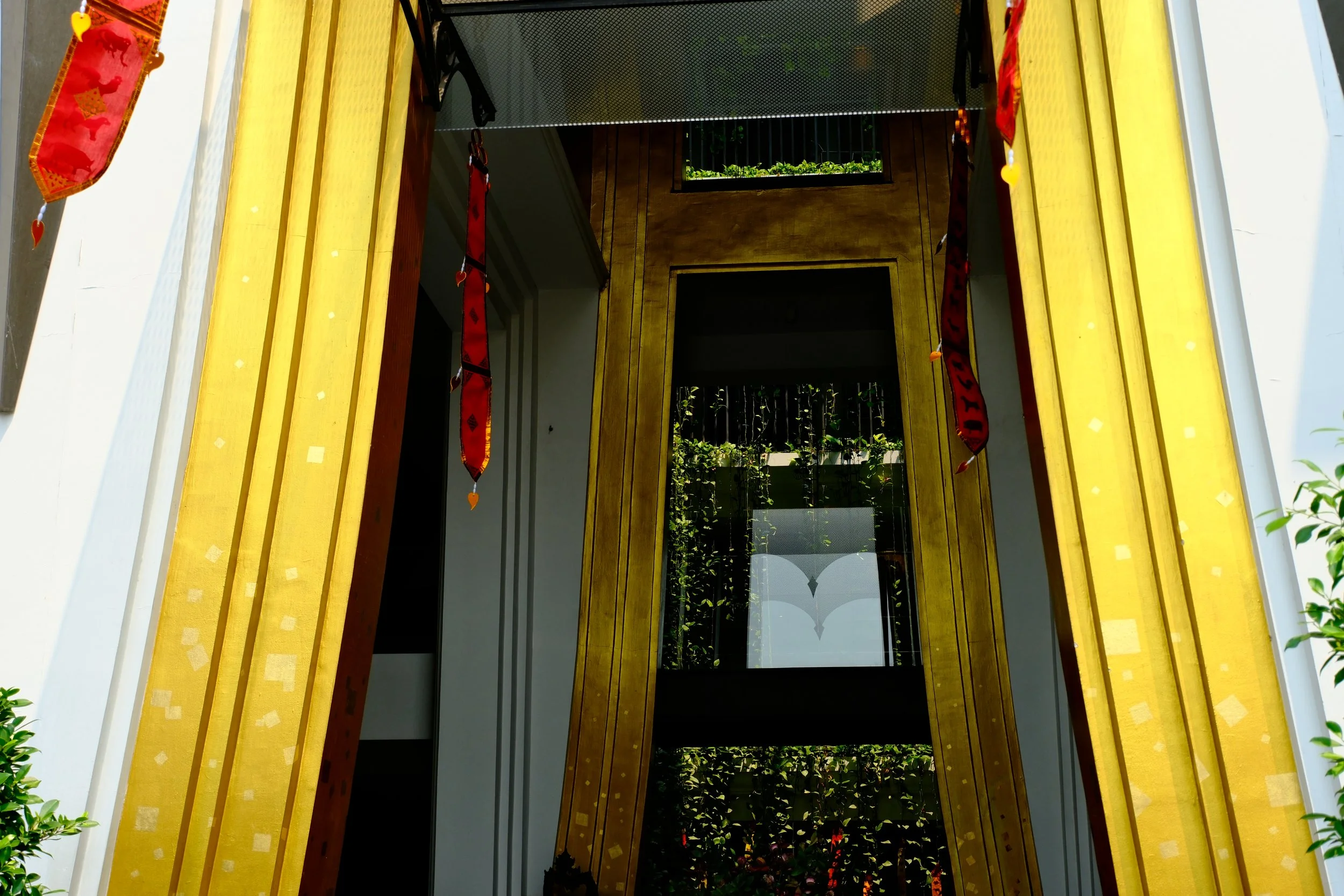 Decorative doorway with golden frame and red hanging ornaments, reflecting greenery and sky in the glass panel.