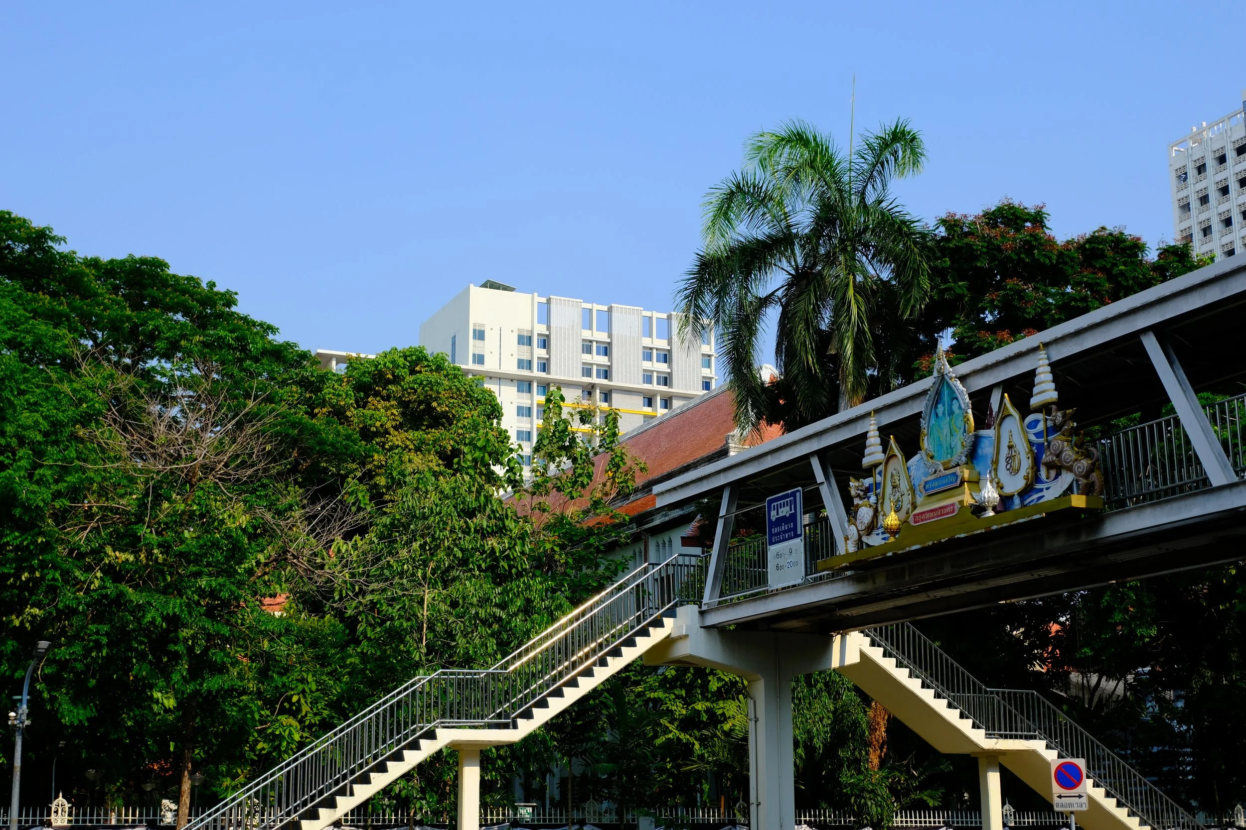 Urban scene with a pedestrian overpass featuring decorative religious statues, lush green trees, and modern high-rise buildings in the background under a clear blue sky.