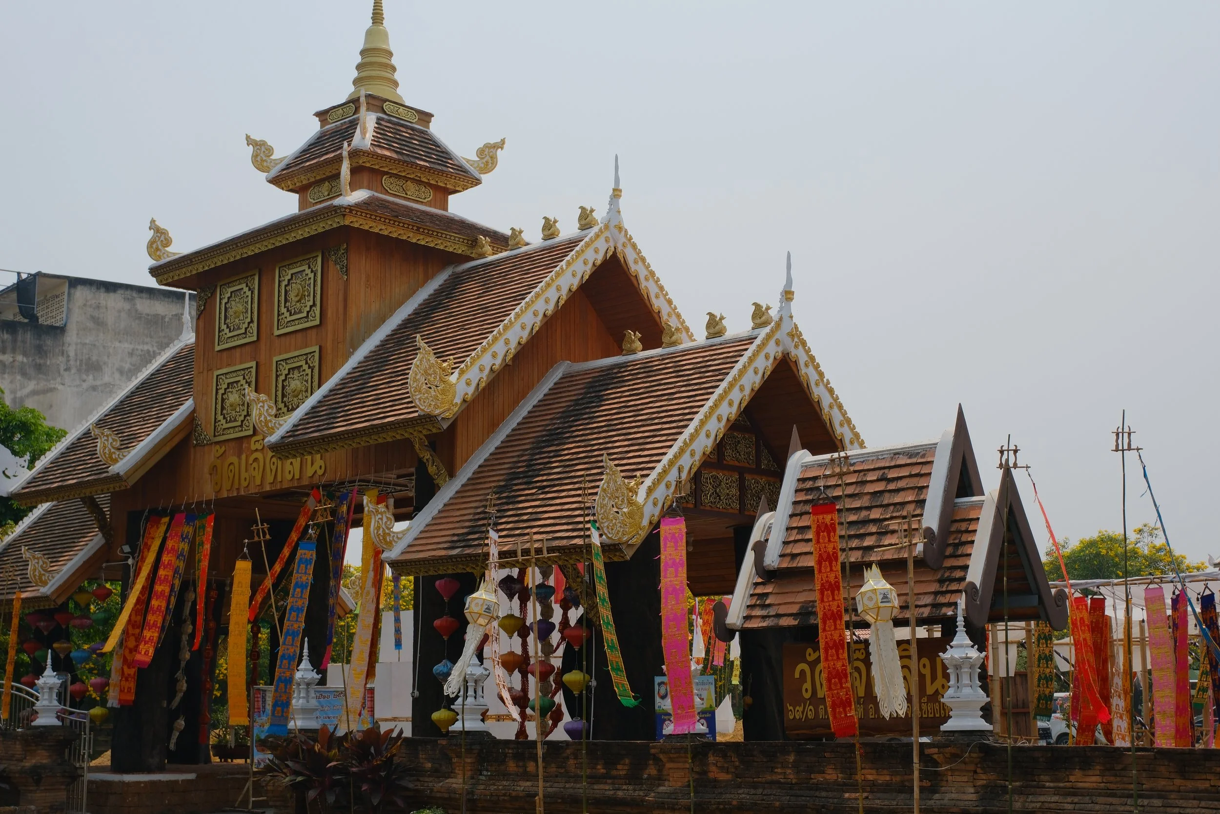 Traditional Thai temple with ornate golden decorations, colorful banners, and hanging lanterns, set against a cloudy sky.