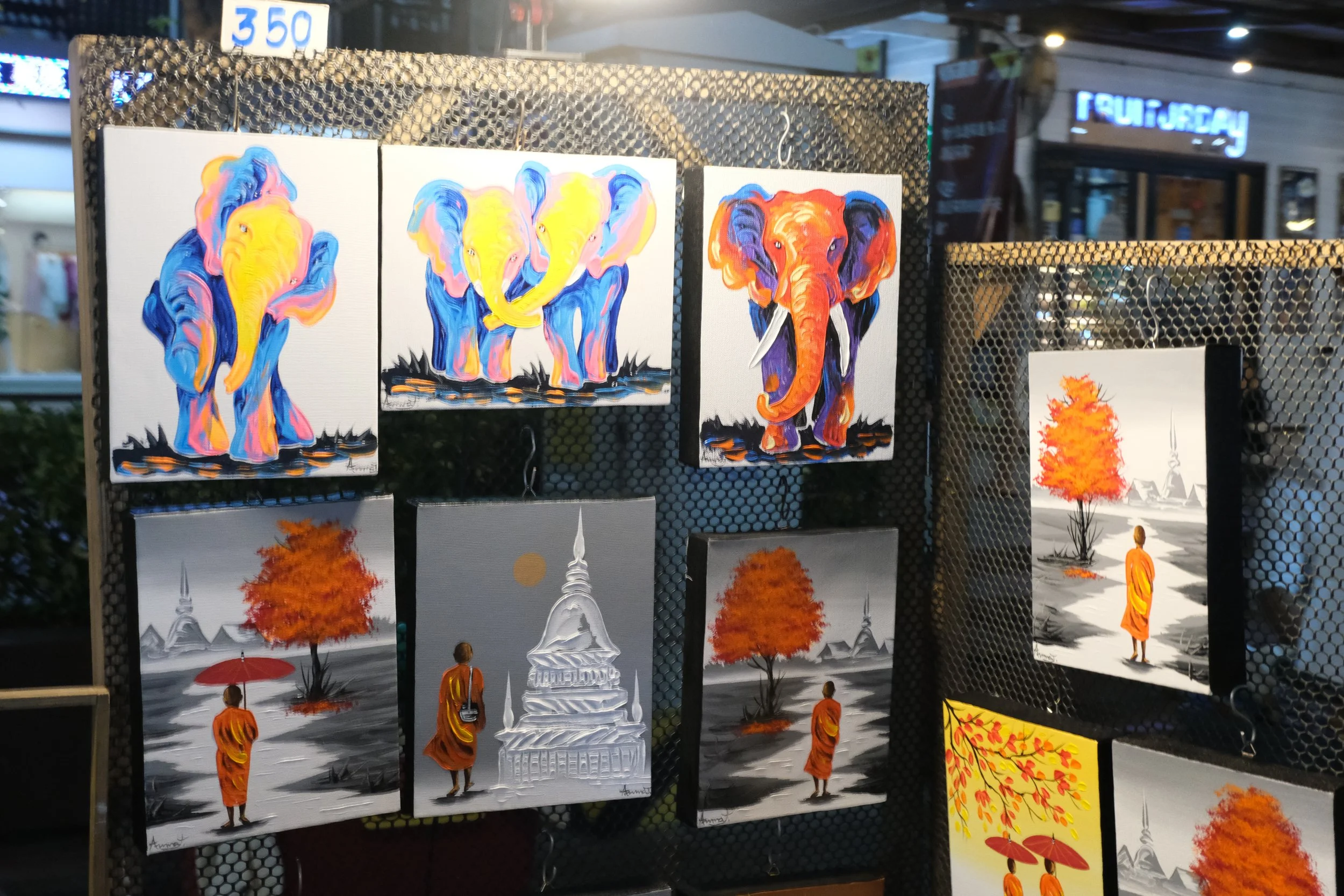 Art display featuring colorful elephant paintings and landscape scenes with a monk, red umbrella, and pagoda.
