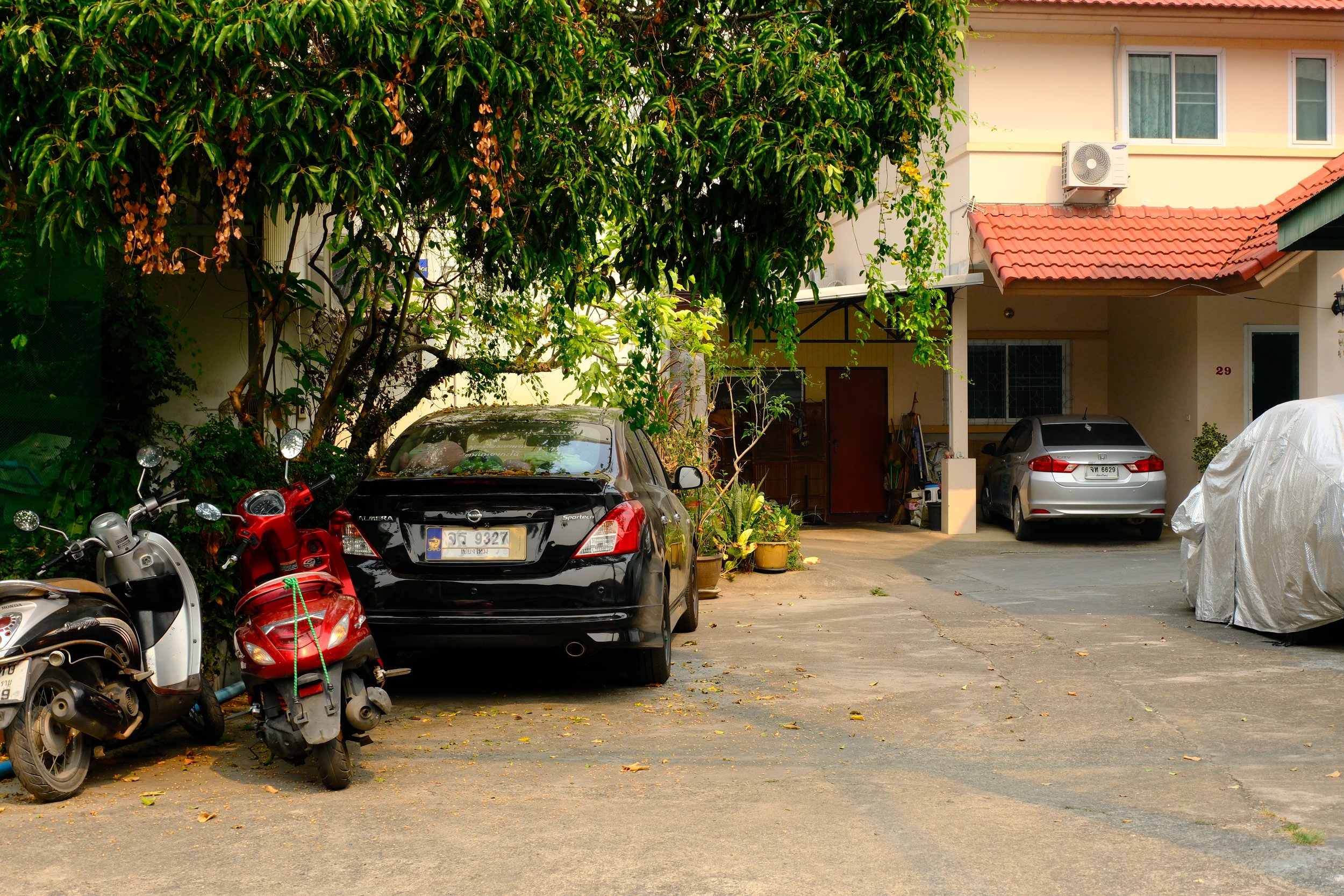A residential driveway with cars and motorbikes parked, some covered with protective sheets, next to a house with potted plants, trees, and an attached garage.