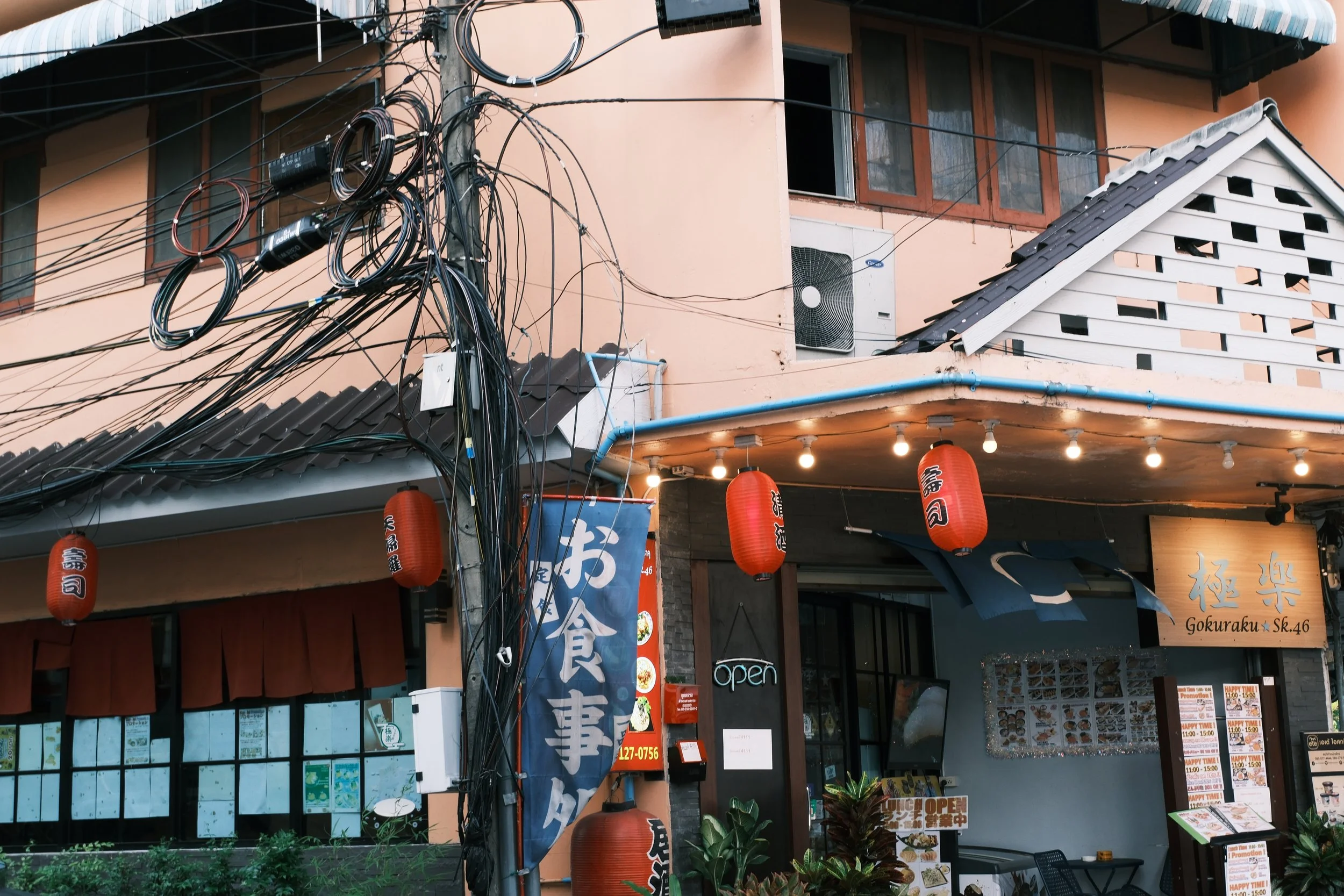 Exterior view of a Japanese restaurant with red lanterns, a blue vertical banner with Japanese characters, and a sign with the restaurant's name 'Gokuraku' in English. There are potted plants outside and the building has a pink facade with windows an