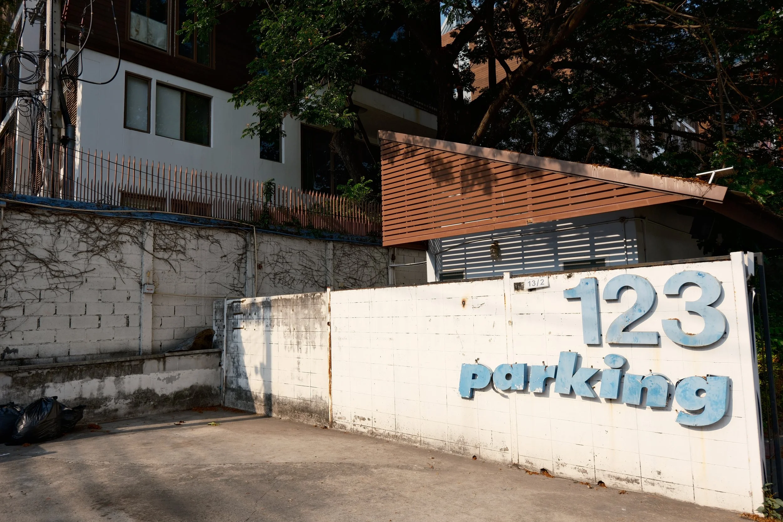 Parking area with a white wall and a sign displaying the address '123' in blue letters and 'parking' written below. Trash bags are on the ground, and a building with windows and a wooden fence are in the background, along with some trees.