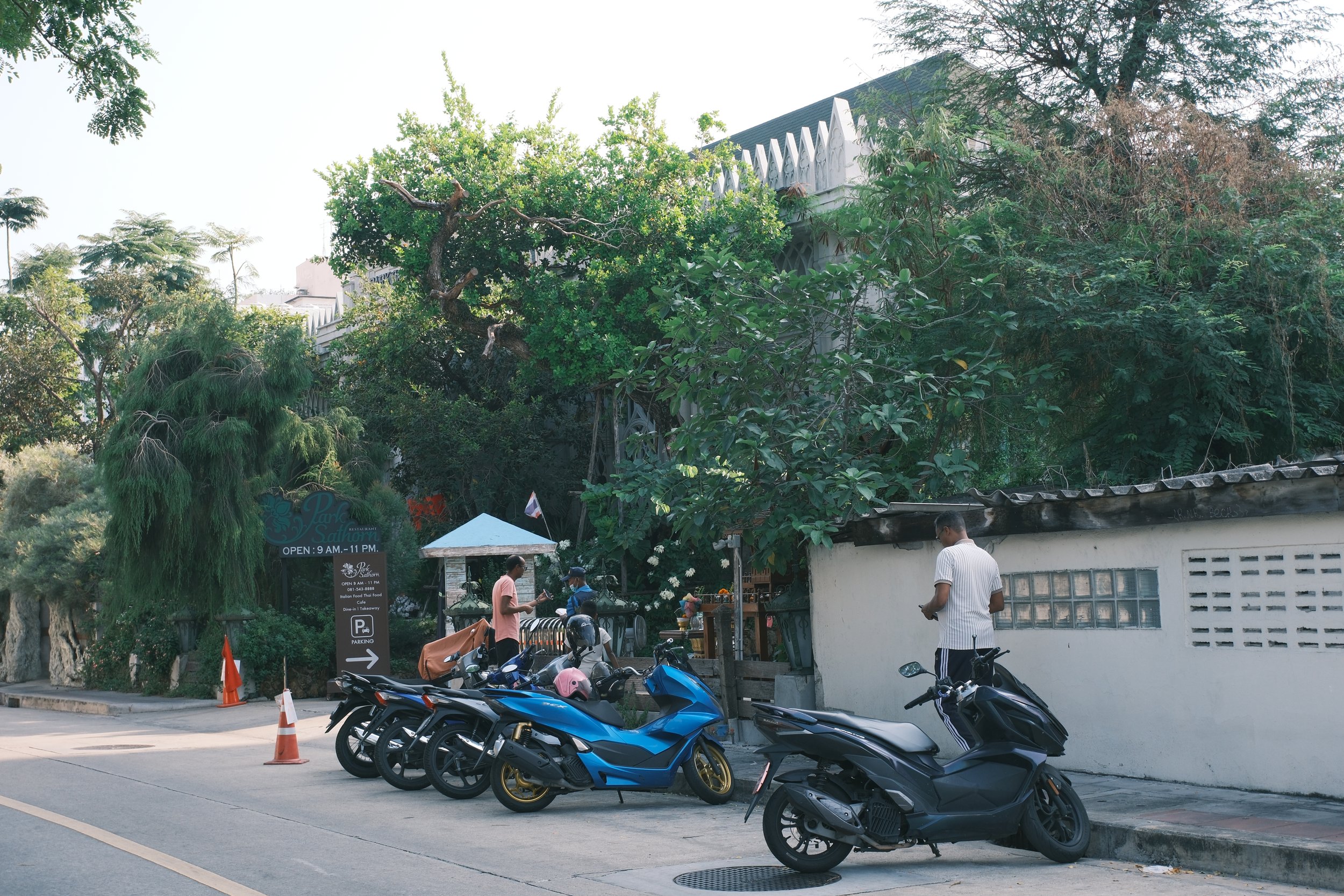 A street scene with several parked motorcycles and scooters near a white wall, with a few people standing and talking nearby. There are trees and a sign for a restaurant in the background.