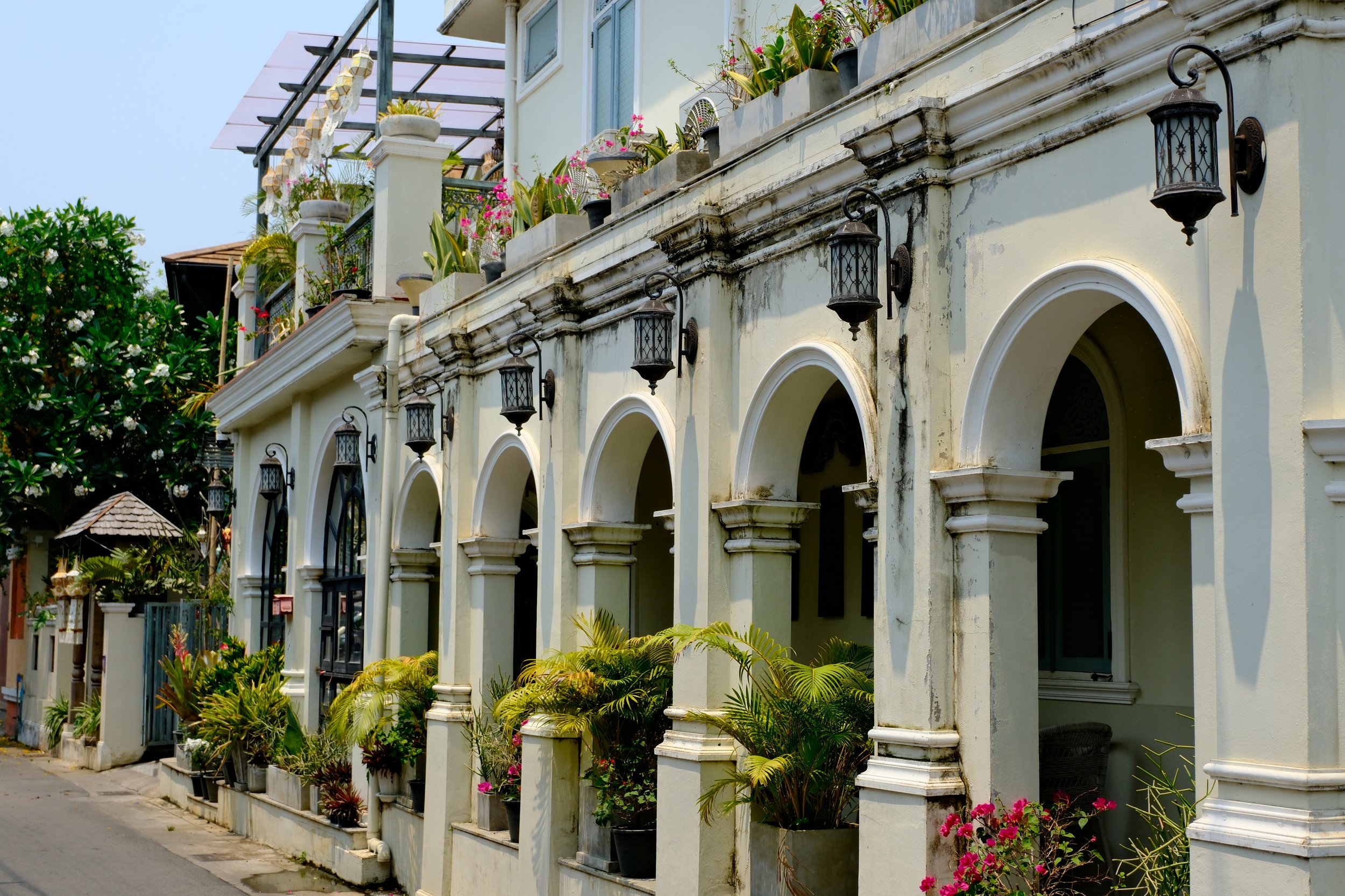 A row of arched doorways and windows on a cream-colored building with hanging lanterns and potted plants on a balcony and on the ground level.
