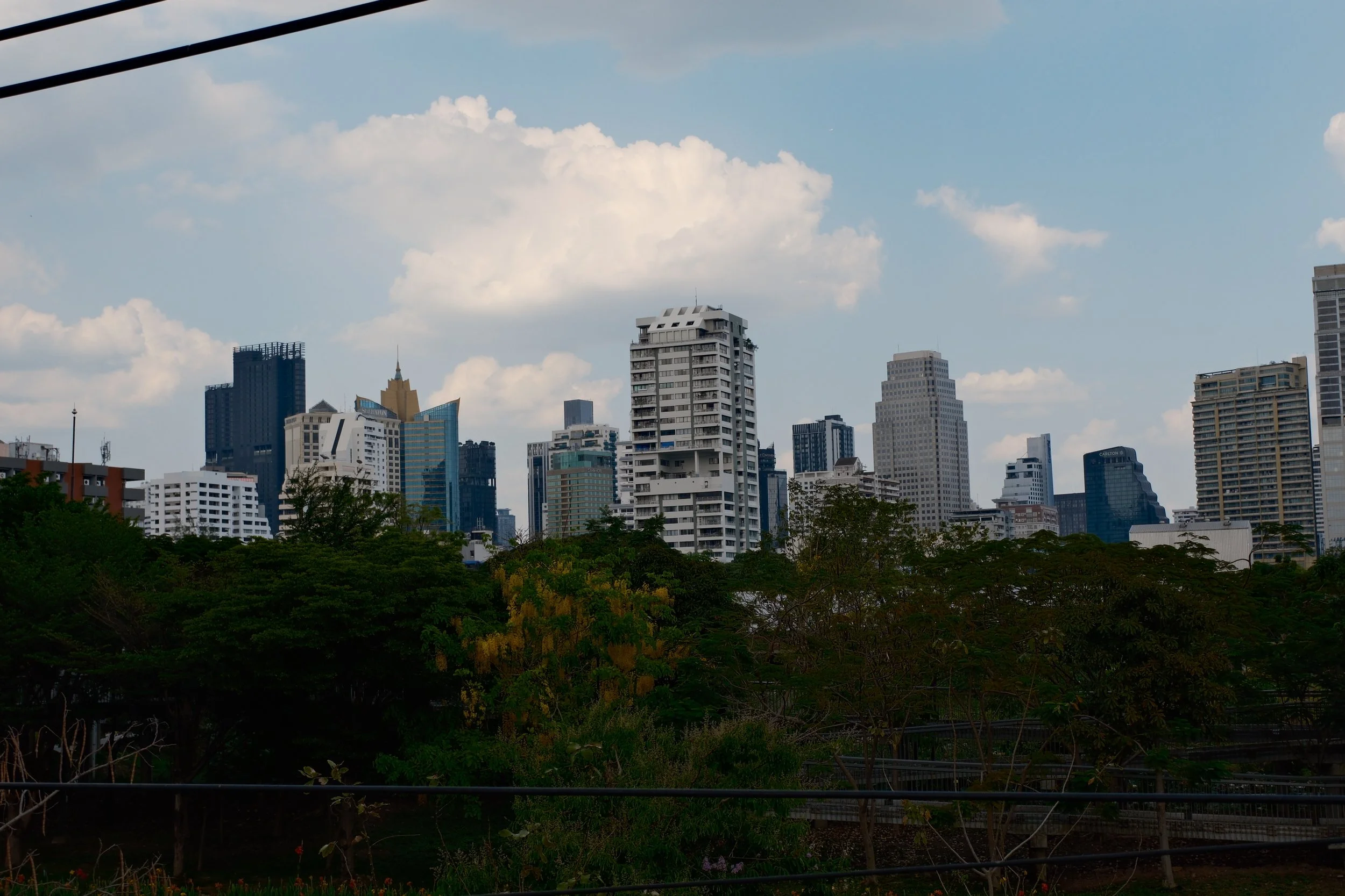 City skyline with tall skyscrapers and a foreground of green trees under a partly cloudy sky.