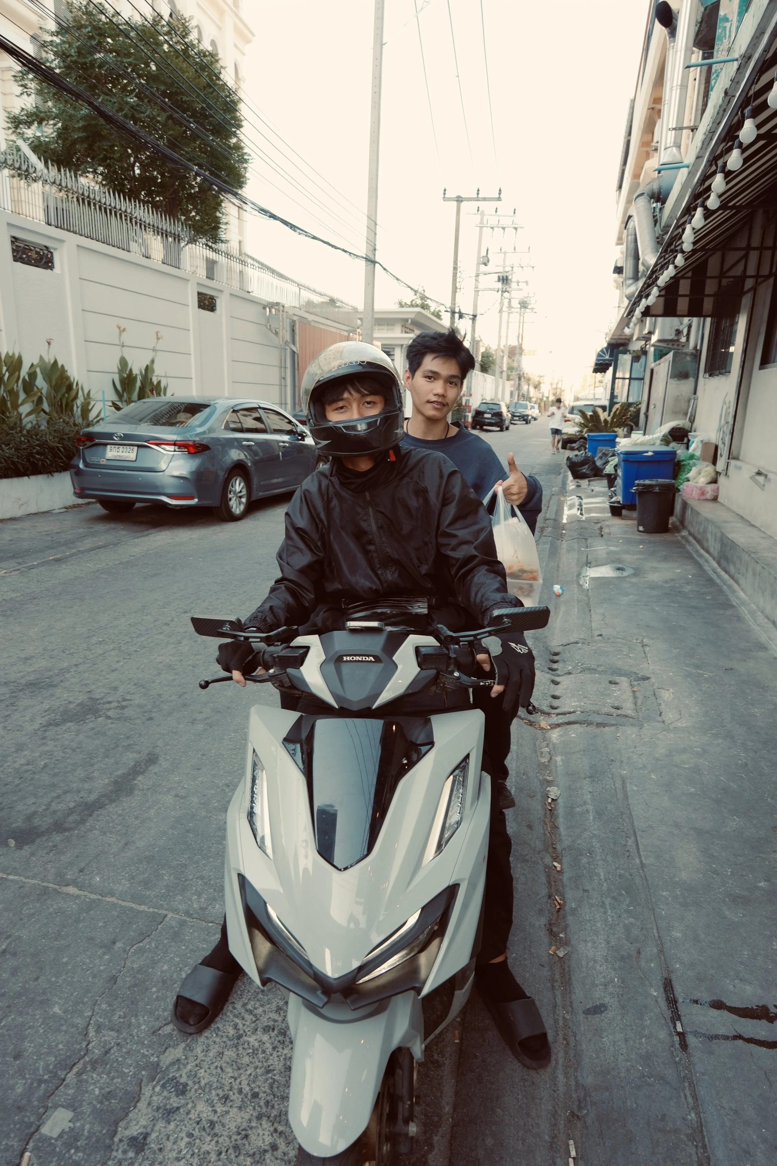 Two young men, one on a motorcycle and the other standing behind, on a city street with parked cars and buildings.