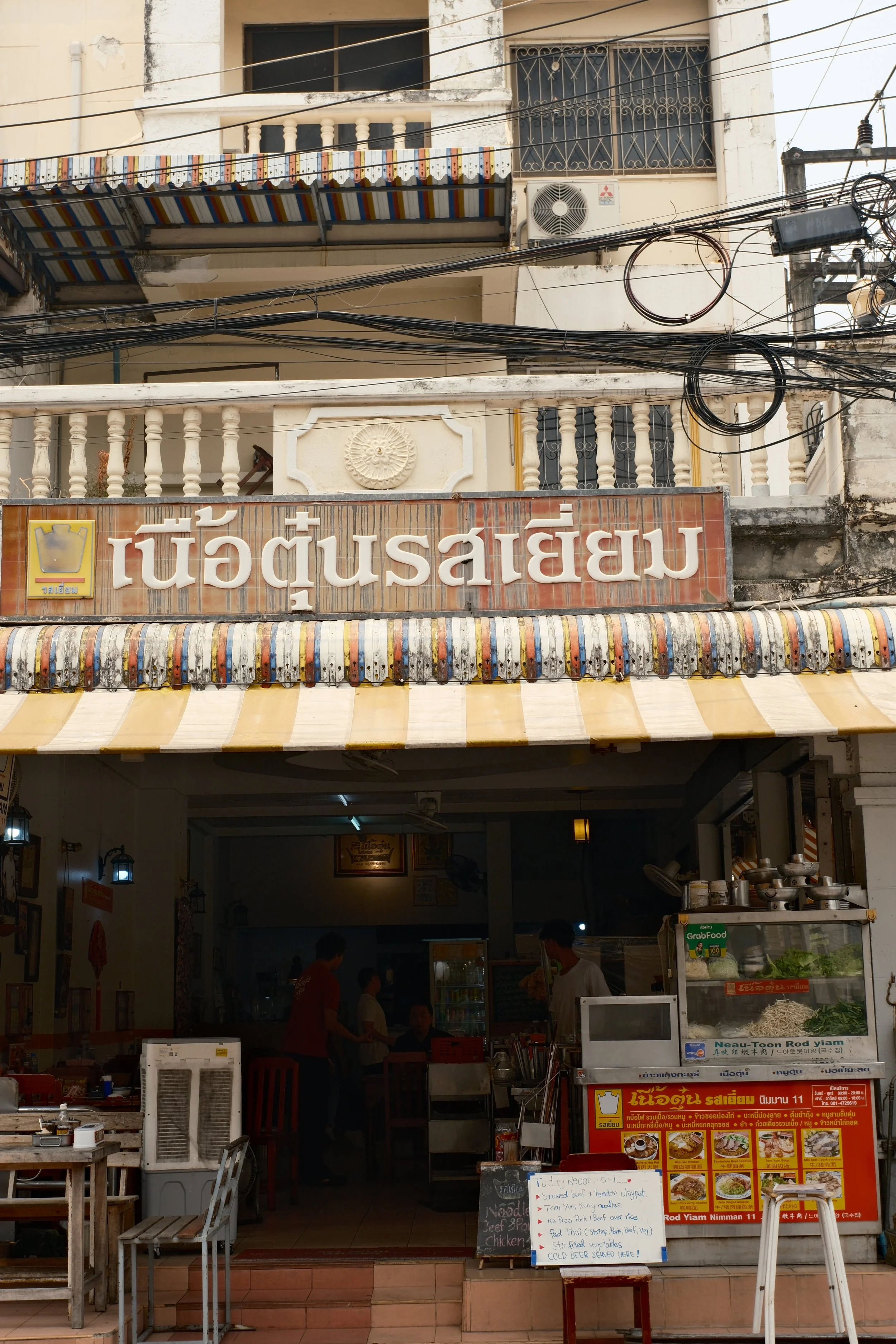 A restaurant with a Thai sign and yellow striped awning, showing the interior with staff and menu items displayed outside.